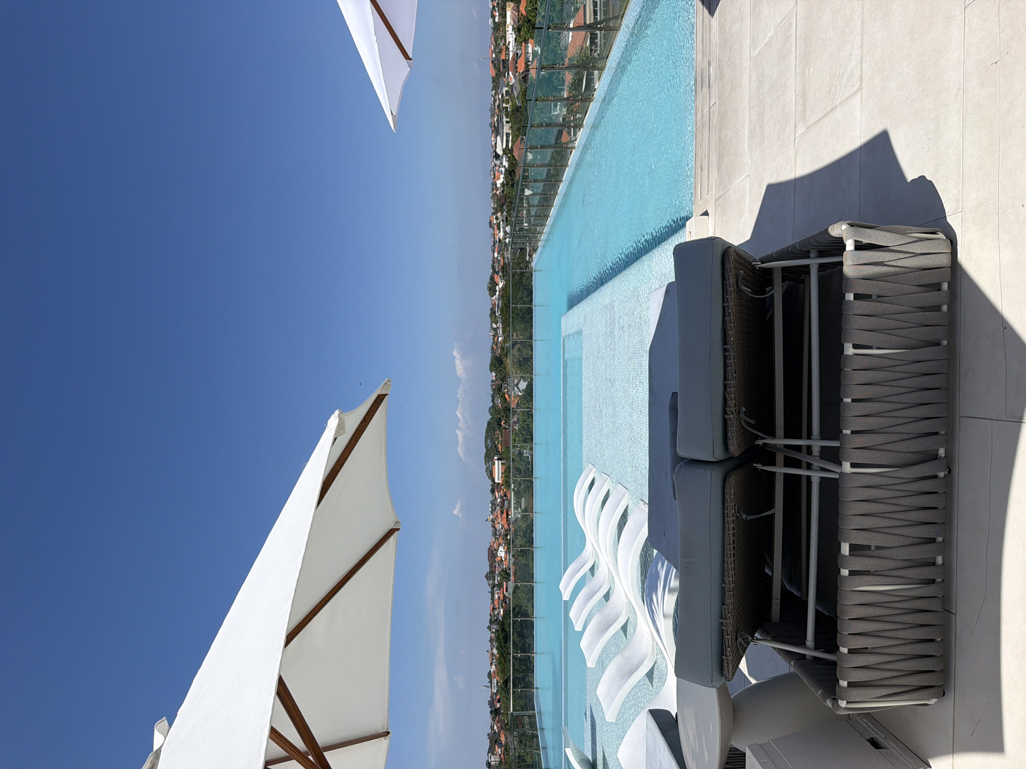This image captures a vibrant, sunny outdoor scene, likely from a high vantage point such as a rooftop or upper-floor terrace overlooking a town. The main subject is a light blue infinity pool, its surface still and reflecting the bright sky. The pool features a shallow area with modern, white, wavy-shaped lounge chairs partially submerged in the water.
To the right of the pool, on light-colored tiled ground, are two grey woven outdoor lounge chairs with light grey cushions, casting strong, defined shadows indicative of bright sunlight. Two large white patio umbrellas with visible wooden support frames are present in the foreground and top left, contrasting against the deep blue, cloudless sky.
The background reveals a sprawling urban landscape of numerous buildings, many with reddish-orange or terracotta-colored rooftops, interspersed with green trees. This residential or commercial area stretches into the distance, suggesting a dense settlement. A faint hint of cloudiness is visible along the distant horizon line to the left.
The scene appears to be set on a clear, bright day, likely late morning or early afternoon, given the intensity and direction of the shadows. There are no people visible, lending a sense of tranquility and an unoccupied atmosphere to the resort-like setting. No visible text can be identified in the image. This serene poolside view is located in North Kuta, Indonesia.