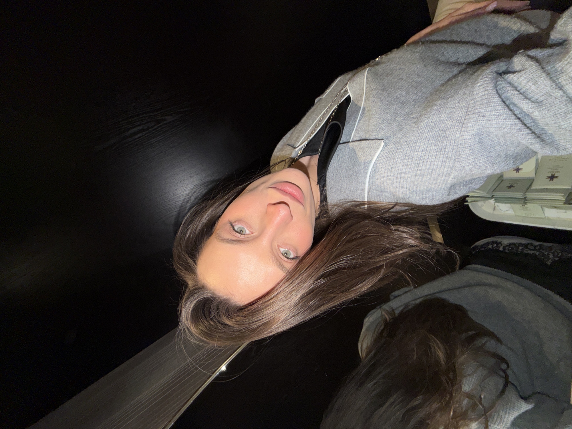 A person is taking a selfie, indoors. The person has long brown hair and light eyes. They are wearing a gray sweater with white trim and a black top. Stacks of small booklets are visible on a tray to the upper right. The background is dark. The location is not discernible from the provided image.
