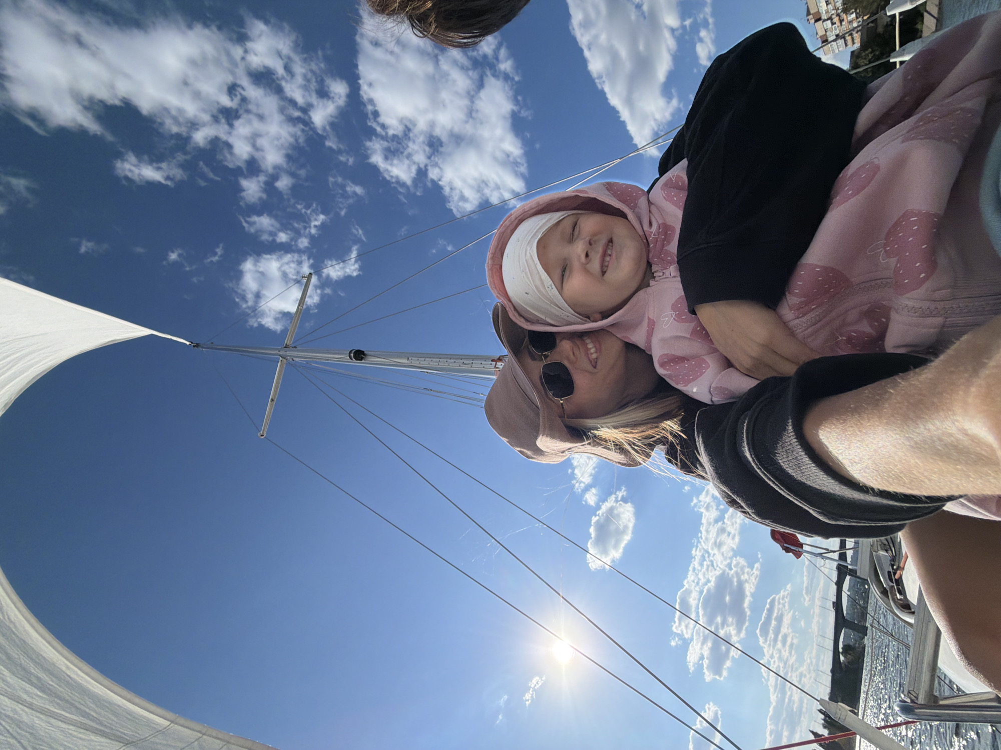 This image captures a mother and child on a sailboat on a sunny day, with a bright blue sky dotted with white clouds overhead. The sailboat's white sail is visible in the upper left of the frame, and the mast and rigging extend upwards. The sun shines brightly, casting a lens flare to the left of the frame.
In the foreground, a woman wearing sunglasses and a light-colored hat smiles as she holds a young child. The child, wearing a pink hoodie adorned with strawberries and a white head covering, is also smiling. They appear to be posing for a selfie.
In the background, a body of water reflects the sky and clouds. A bridge with several arches can be seen in the distance, spanning the water. Along the far bank of the water, buildings are visible, suggesting an urban or semi-urban environment.
The scene is consistent with being on the Volga River in the city of Kamyshin, Russia. The bridge in the background is likely the Kamyshin Bridge, a road bridge that crosses the Volga River. The presence of a sailboat on the river also indicates a location within Kamyshin.