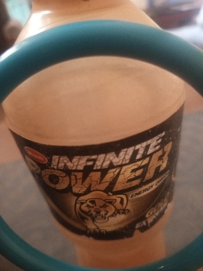 A bottle labeled "Infinite Power" energy drink is framed by a blue, circular object. The energy drink label is black and gold, featuring a cougar graphic. The "Power" text is cracked, suggesting strength. The bottle's background is a light beige or tan.
