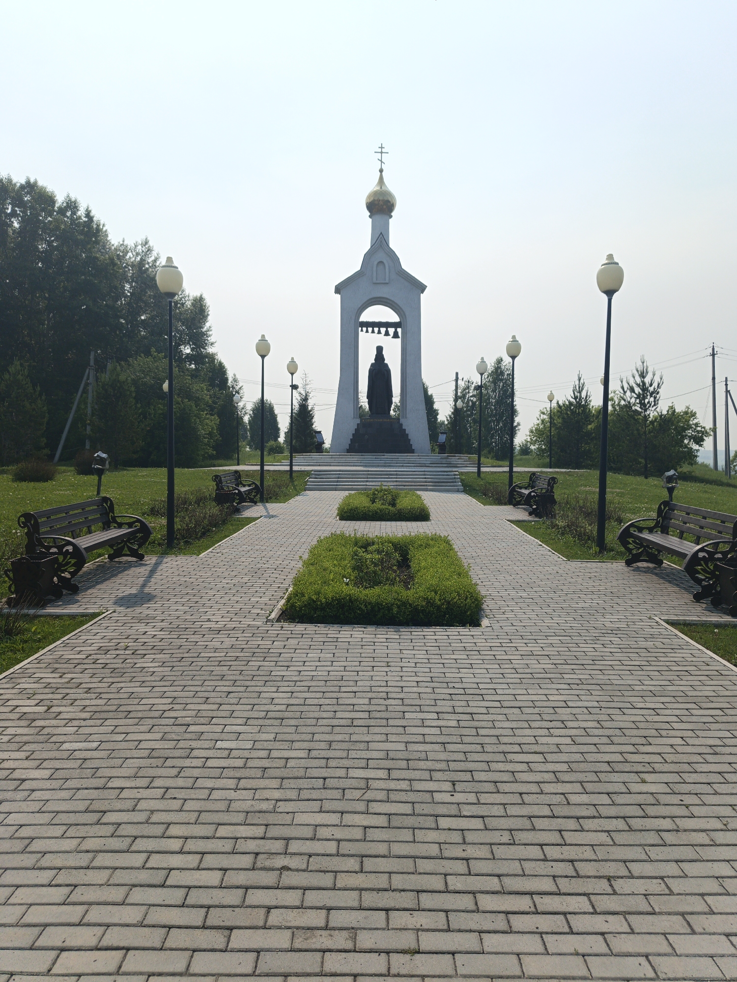 The image shows a memorial chapel with a statue of a person in front of it. The chapel is white with a gold dome. The statue is dark in color and stands on a pedestal. In front of the chapel, there is a paved walkway lined with benches and lampposts. The walkway leads away from the viewer towards the chapel. The area is grassy with trees in the background. The sky is hazy and light blue. The location is identified as Kalachevo, Russia.