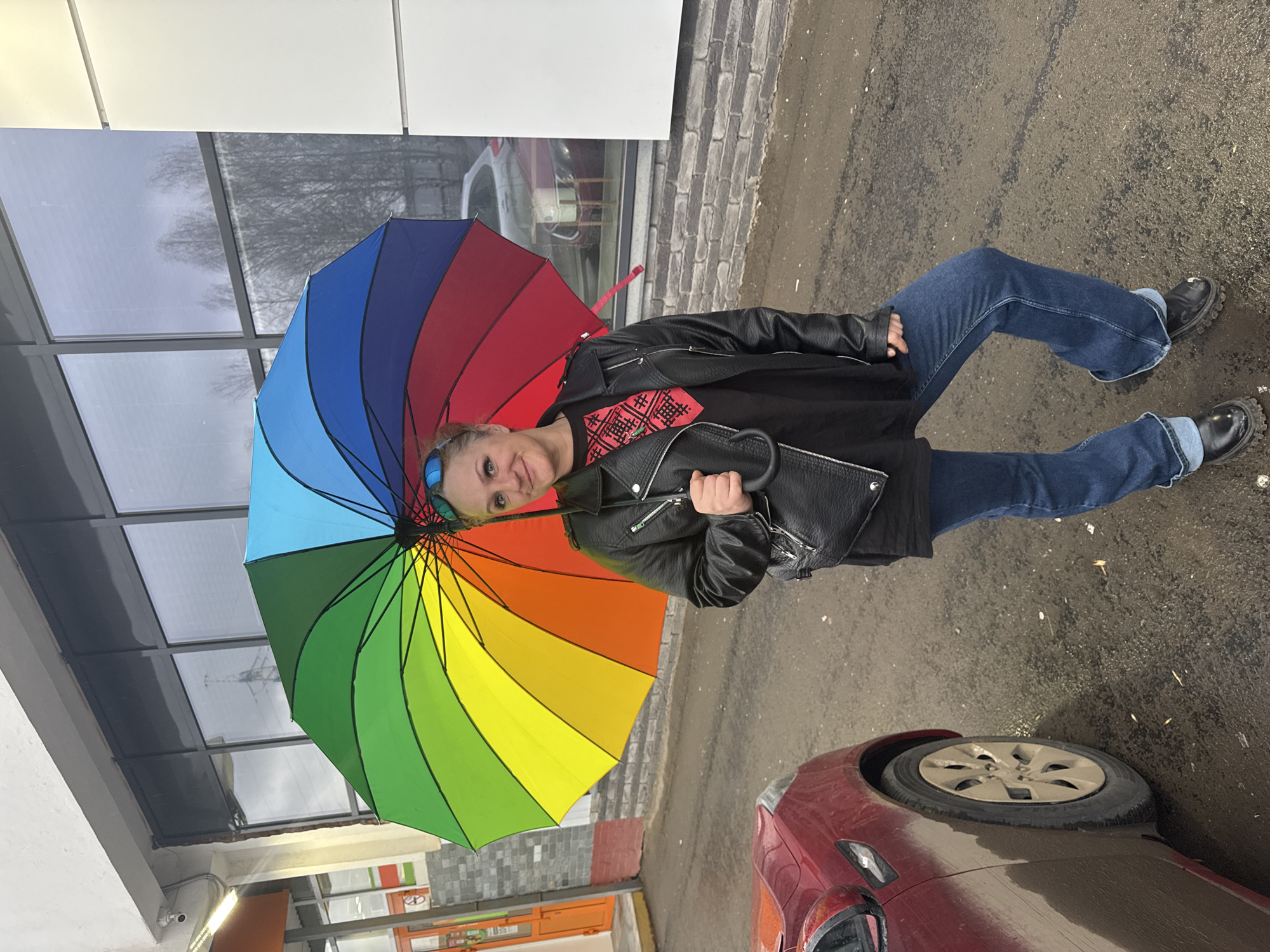 A woman in jeans and a black leather jacket stands outside holding a rainbow umbrella. She is smiling and facing the camera. She is standing on a wet sidewalk next to a building and a red car. The building has a modern glass facade. In the background, there is a reflection of trees in the glass. The location appears to be an urban environment, likely in Izhevsk, Russia.