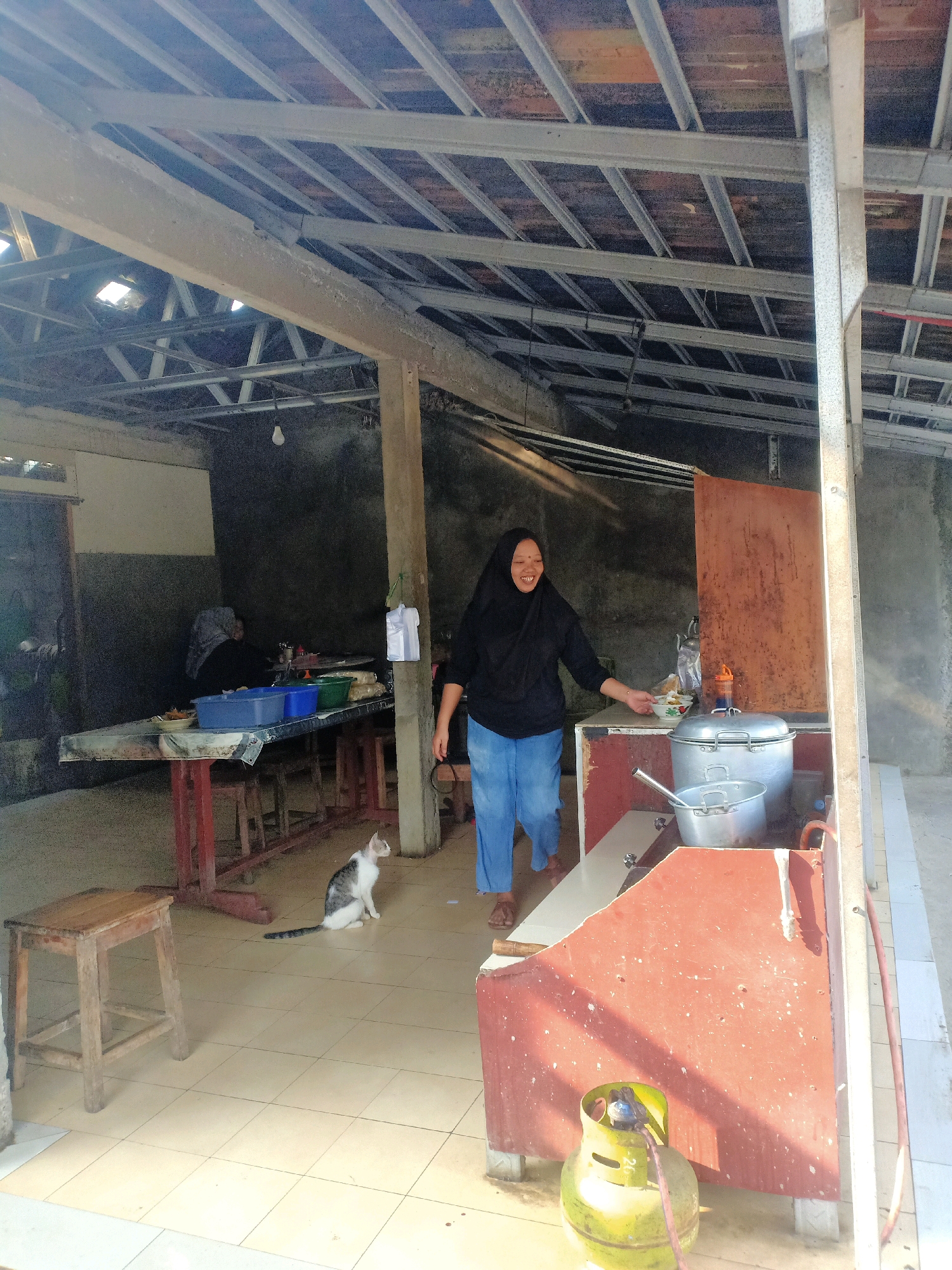 In Jatinom, Indonesia, a woman wearing a black hijab and blue pants is standing in a covered outdoor food stall. She is smiling and reaching towards a table. In the background, another person wearing a hijab is seated at a table. A cat is sitting on the tiled floor near a wooden stool. The stall has a metal roof structure and concrete walls. Cooking equipment, including a large pot and a gas cylinder, are visible on a red counter. The scene appears to be a typical day at a local eatery.