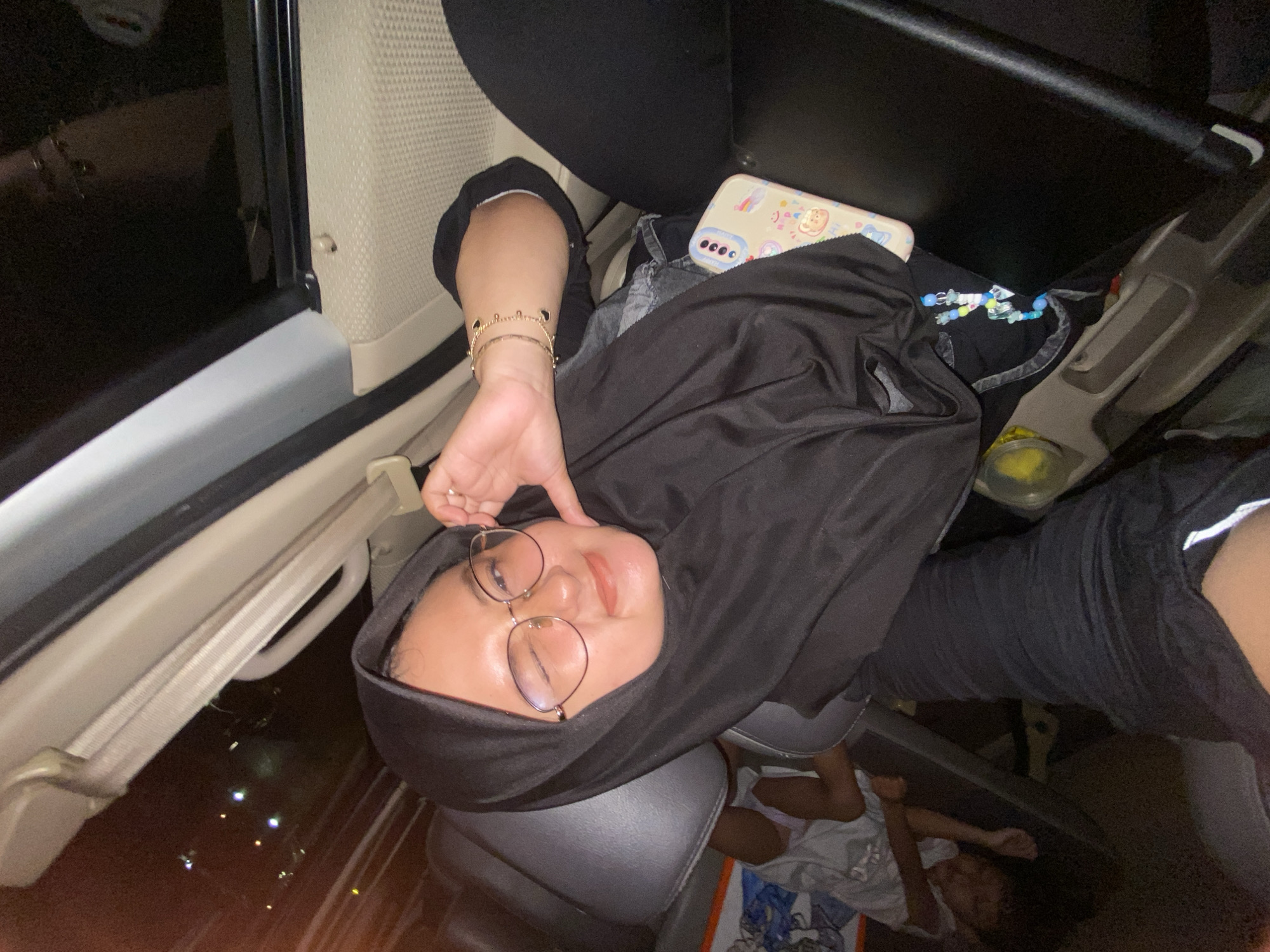 A person wearing glasses and a black hijab is seated in a car. The car door is open. A mobile phone with a decorated case is visible. Outside the car window, it appears to be nighttime with some scattered lights. Another person is lying down in the back of the car. It is not possible to verify the exact location in Surabaya, Indonesia, without additional information.