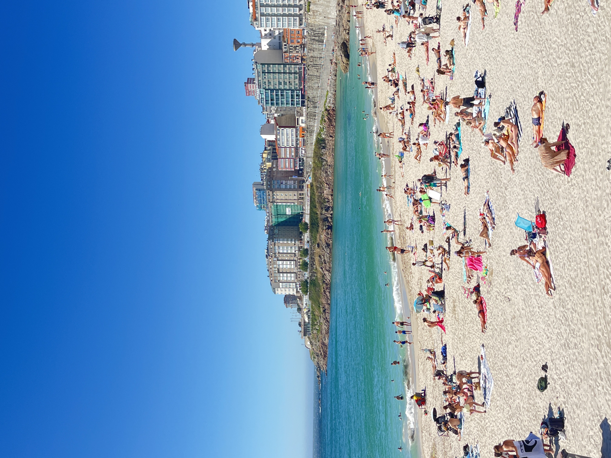 The media file shows a sunny day at a crowded beach in A Coruña, Spain. The scene is a wide shot looking down at a sandy beach filled with people sunbathing and swimming in the clear, turquoise ocean. The water is calm with small waves gently lapping the shore. Buildings line the edge of the beach, with a rocky outcrop separating the sand from the urban landscape. The sky is a clear, bright blue, indicating good weather and likely midday. Many people are scattered across the sand, some lying on towels, others sitting up, and some wading in the water. There are various beach accessories visible, including umbrellas, towels, and bags. The overall impression is one of a popular summer day at a coastal city.