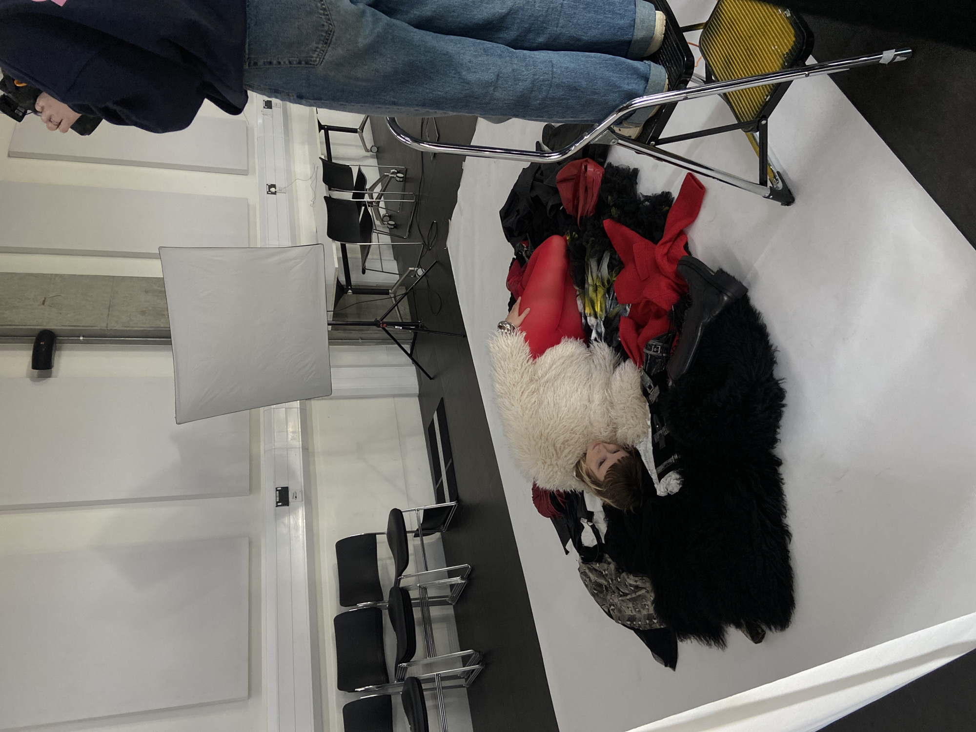 A photograph shows a person lying in a pile of clothes on a white surface. The person is wearing a white fur coat and red tights. There are also red, black, and camouflage clothes in the pile. A person stands over the pile with a camera. There is a black and yellow chair and a lighting rig to the right of the pile of clothes. There are several black chairs and acoustic panels on the wall in the background. The location appears to be a photo studio. There is no clear indication of the city of London, United Kingdom.