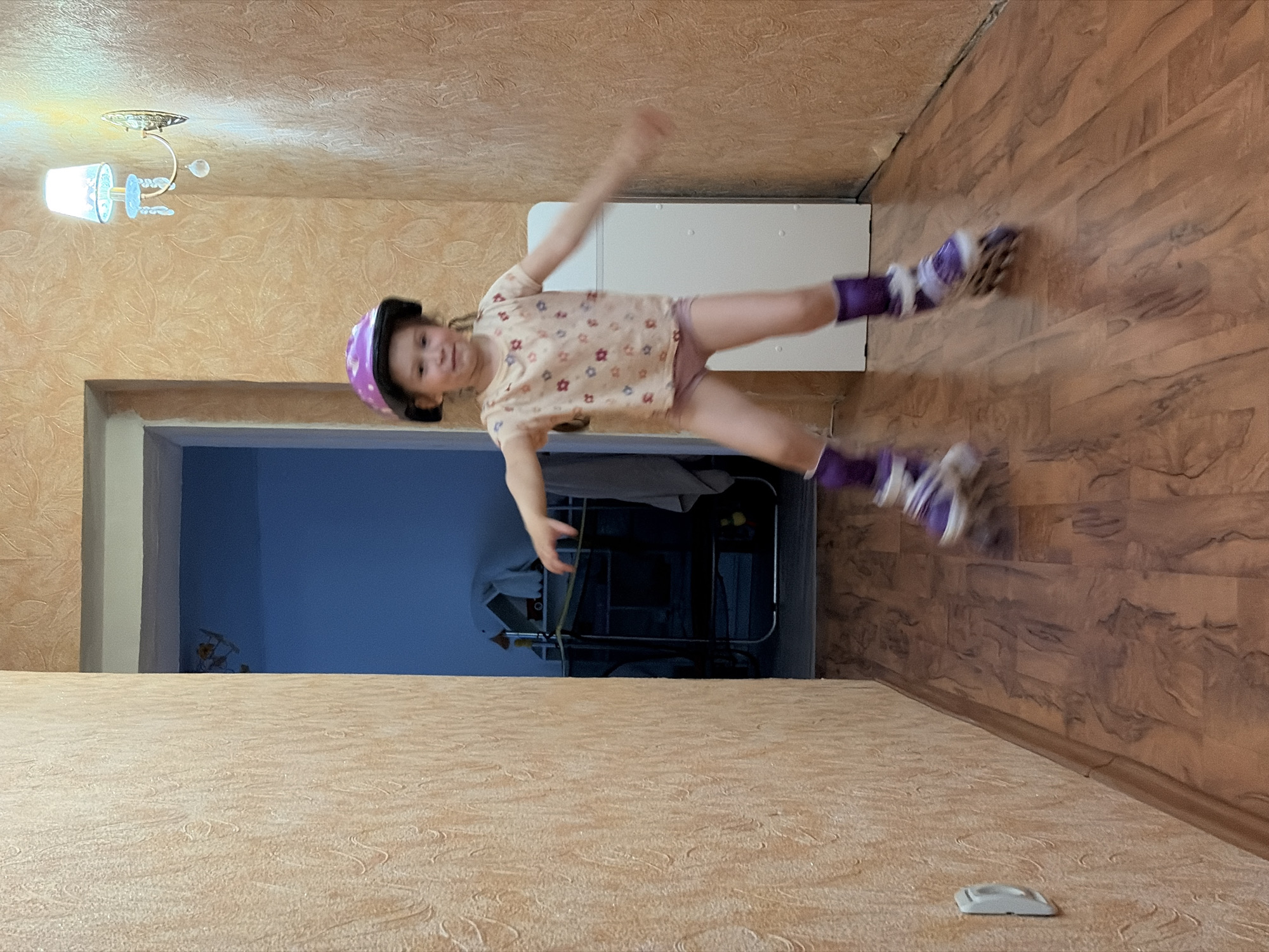 A young girl wearing roller skates and a helmet is suspended horizontally in mid-air, appearing to be jumping or performing a stunt. She is wearing a light-colored shirt with a floral pattern and purple roller skates and socks. The scene takes place indoors, in a room with floral wallpaper on the upper part of the walls and wood-patterned wallpaper on the lower part. A doorway is visible in the background, leading to a room with blue walls and containing a metal shelving unit. A light fixture is affixed to the ceiling. The photograph was taken in what is likely a residential setting in Chaikovsky, Russia, based on the user's request.