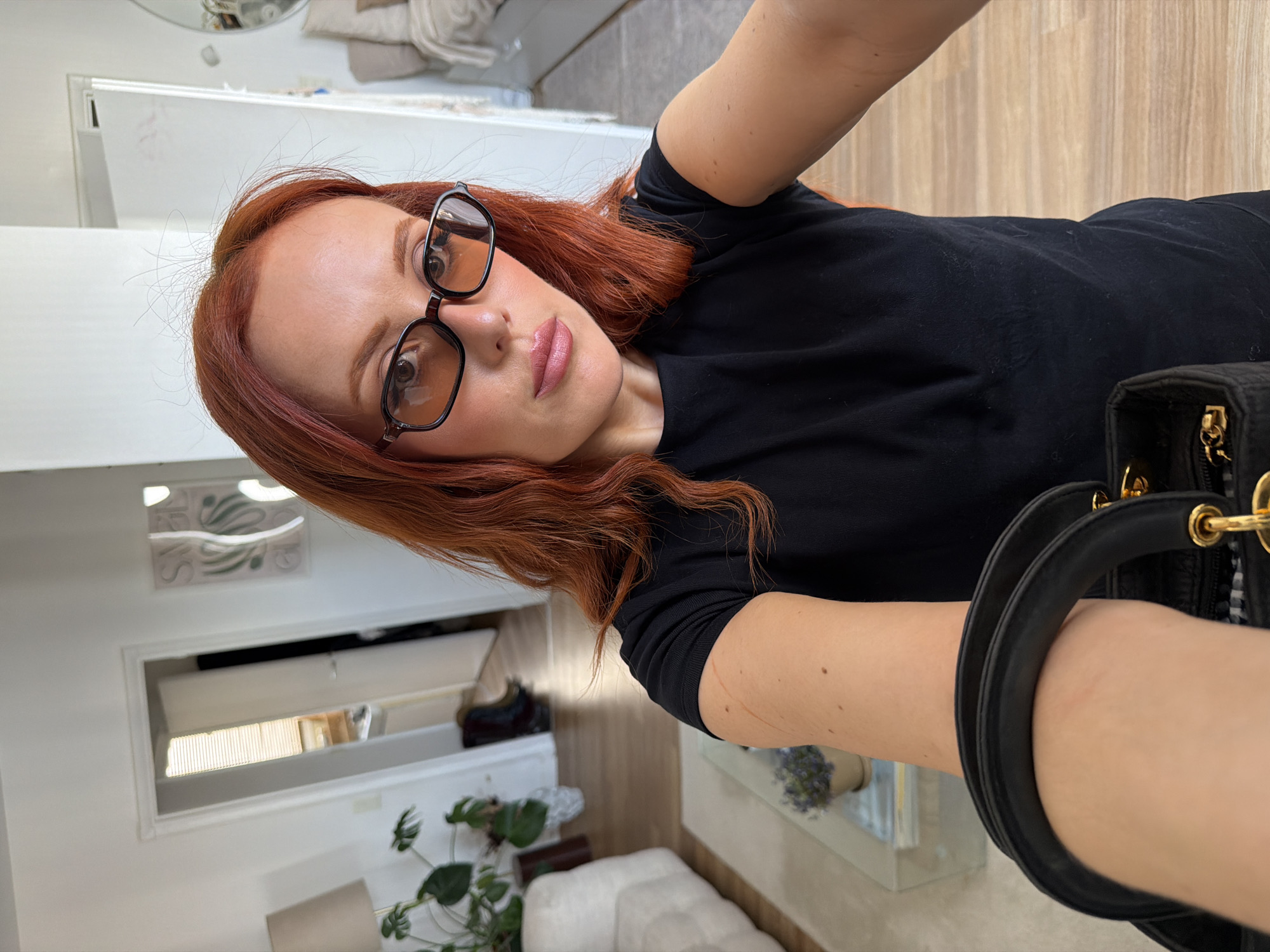 A red-haired woman wearing glasses and a black shirt is taking a selfie in what appears to be her home. A black handbag with gold hardware is partially visible in the foreground. The background includes a white wall with artwork, a window, a plant, and light-colored furniture. The scene does not depict Paris, France.