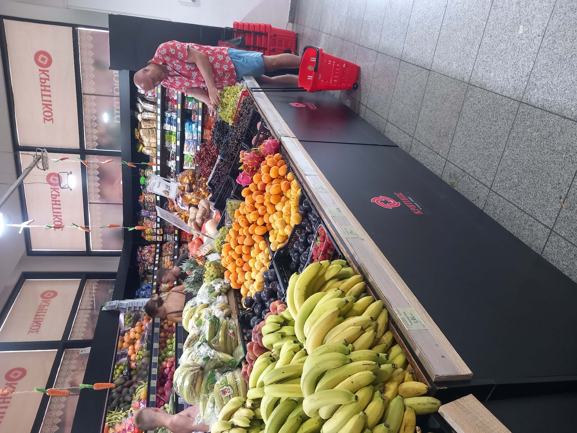 In the city of Kos, Greece, a man in a red floral shirt and blue shorts is shopping at a supermarket, selecting green grapes from a display. He stands near a selection of fruits, including bananas, apricots, plums, and dragon fruit, as well as rows of packaged goods. Two other shoppers, a man and a woman, are visible in the background, also browsing the produce. The supermarket's interior features tile flooring, illuminated shelves, and signage. The scene suggests a typical grocery shopping experience.