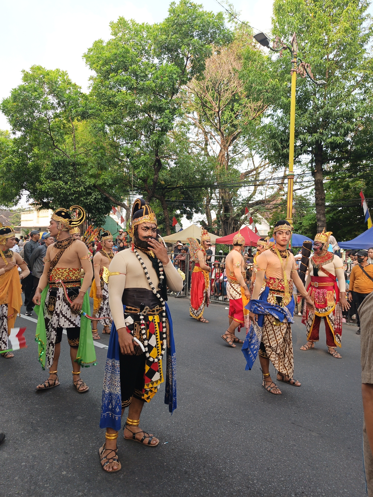 A group of men in traditional Javanese attire, likely a cultural parade or performance, are walking on a paved street in Klaten, Indonesia. The men are adorned in ornate headdresses, elaborate necklaces, loincloths, and patterned sarongs. Some wear gold armbands and anklets. One man in the foreground has a dark beard and is touching his chin. In the background, there are onlookers, trees, a yellow lamppost, and tents, suggesting a public event. The time of day appears to be afternoon, given the lighting and the shadows.