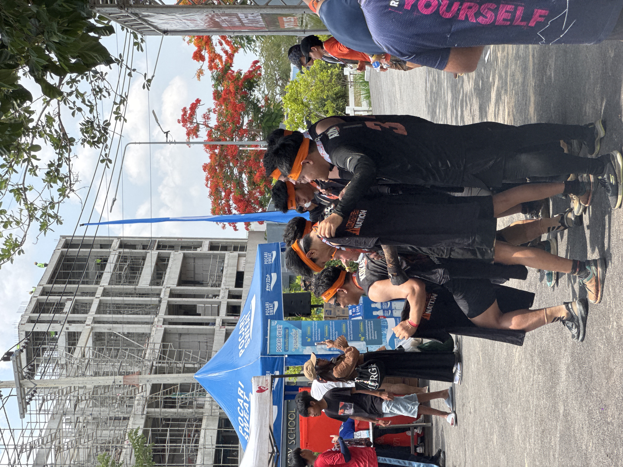 The image shows a group of people, likely participants in an event, gathered near a Pocari Sweat tent in บ้านสำนักตะแบก, Thailand. They are wearing black shirts with orange headbands. The event seems to be a "Tough Mudder" race, as indicated by the branding on the shirts. Some individuals are looking at their phones. In the background, there is a building under construction and a tree with red flowers. There are utility poles and overhead wires. Other individuals are seen in the background. The overall setting appears to be an outdoor event.