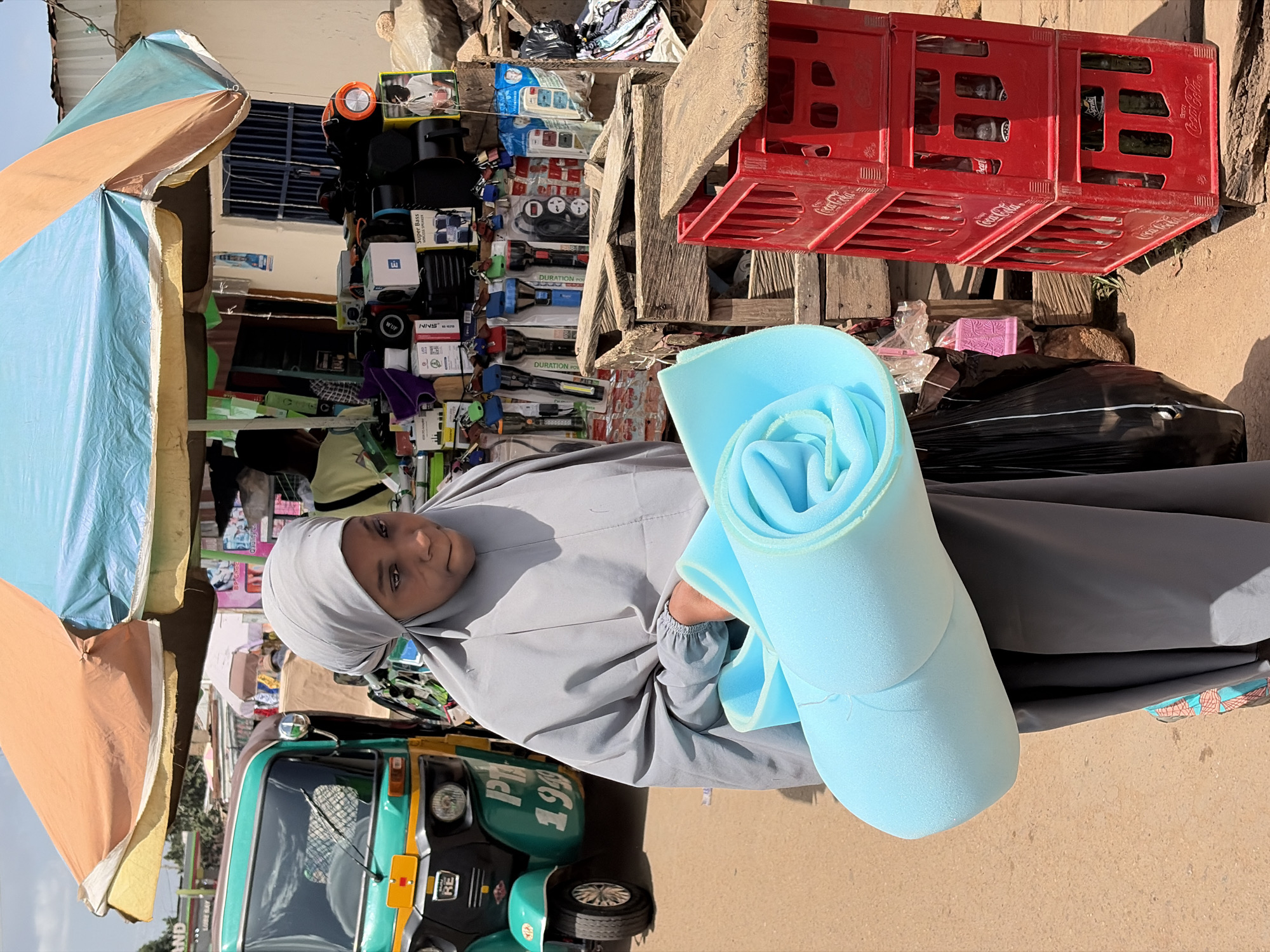A young woman stands in what appears to be an outdoor market in Jos, Nigeria, looking directly at the camera with a neutral to slightly pleasant expression. She is dressed in a light grey abaya and matching hijab. In her arms, she holds a large, rolled-up piece of light blue foam.
Behind her, a vibrant market scene unfolds under bright daylight. To her left, the front of a green and yellow tricycle, commonly known as a keke napep, is visible, with partial text including "IPT" and "199" on its side. Above and behind her, numerous consumer goods are displayed on shelves, including electronics like power strips, flashlights (some labeled "DURATION POWER"), and speakers (with visible text like "Super Bass", "NNS", and "WUP" on their packaging).
To her right, a stack of red plastic Coca-Cola crates is prominent, containing bottles and clearly displaying the "Coca-Cola" logo. Below these crates, a black trash bag and other debris are visible. Overhead, large, colorful market umbrellas in shades of blue, orange, and tan provide shade, indicating a sunny outdoor setting.