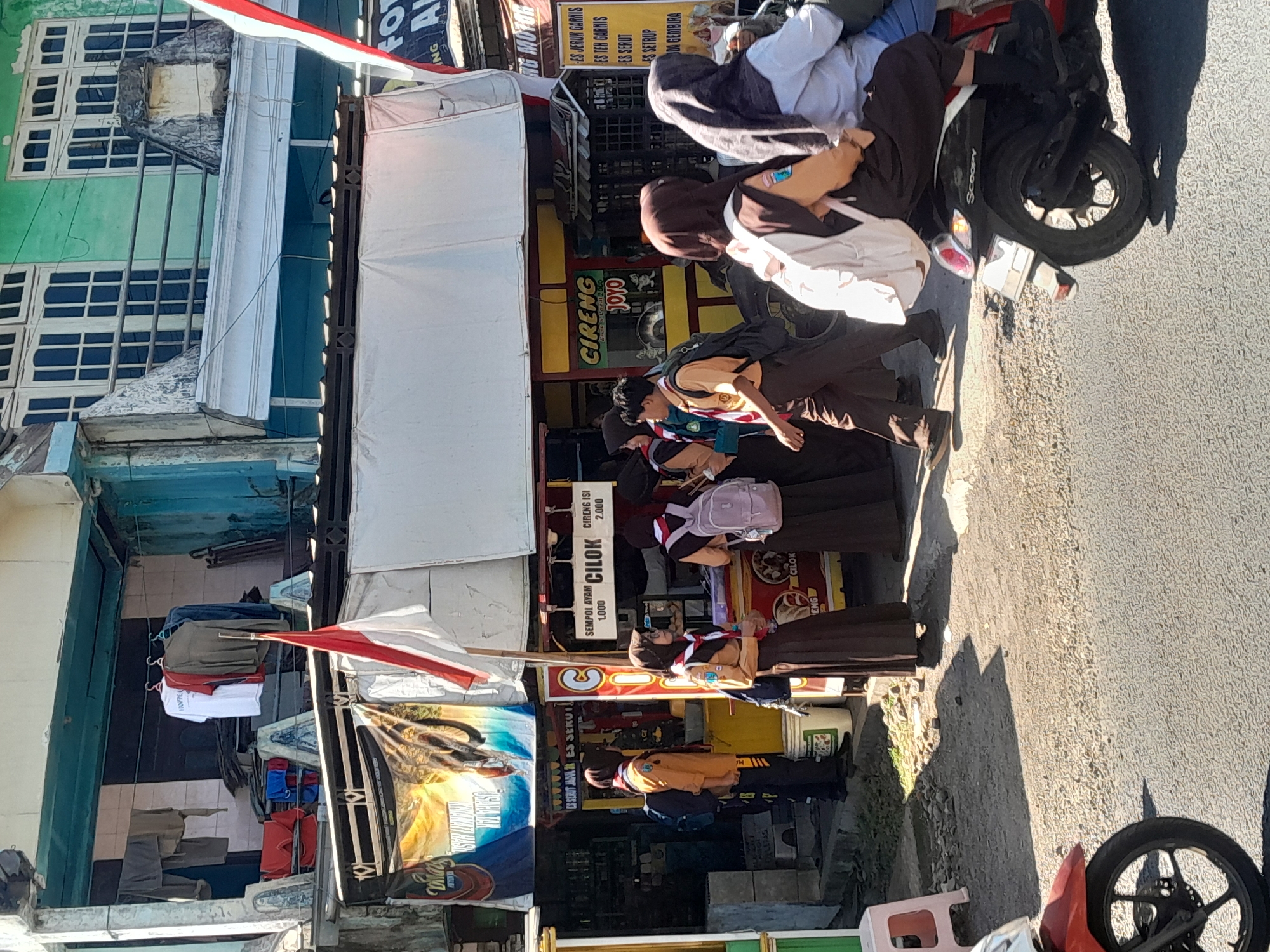 In Srono, Indonesia, a street scene unfolds under bright sunlight. In the foreground, the front wheel and portion of a red scooter are visible. Behind it, a group of students in school uniforms are gathered near a food stall. The students, some wearing hijabs, have backpacks and appear to be either waiting to order or observing the stall. The food stall displays prices and names of items like "Cilok" and "Cireng." Further back, laundry is hung on a line above the stall, with clothing items like shirts and pants visible. The buildings have a mix of architectural styles and colors, including green and teal facades. A red and white Indonesian flag is prominently displayed. The ground is a mixture of asphalt and gravel.