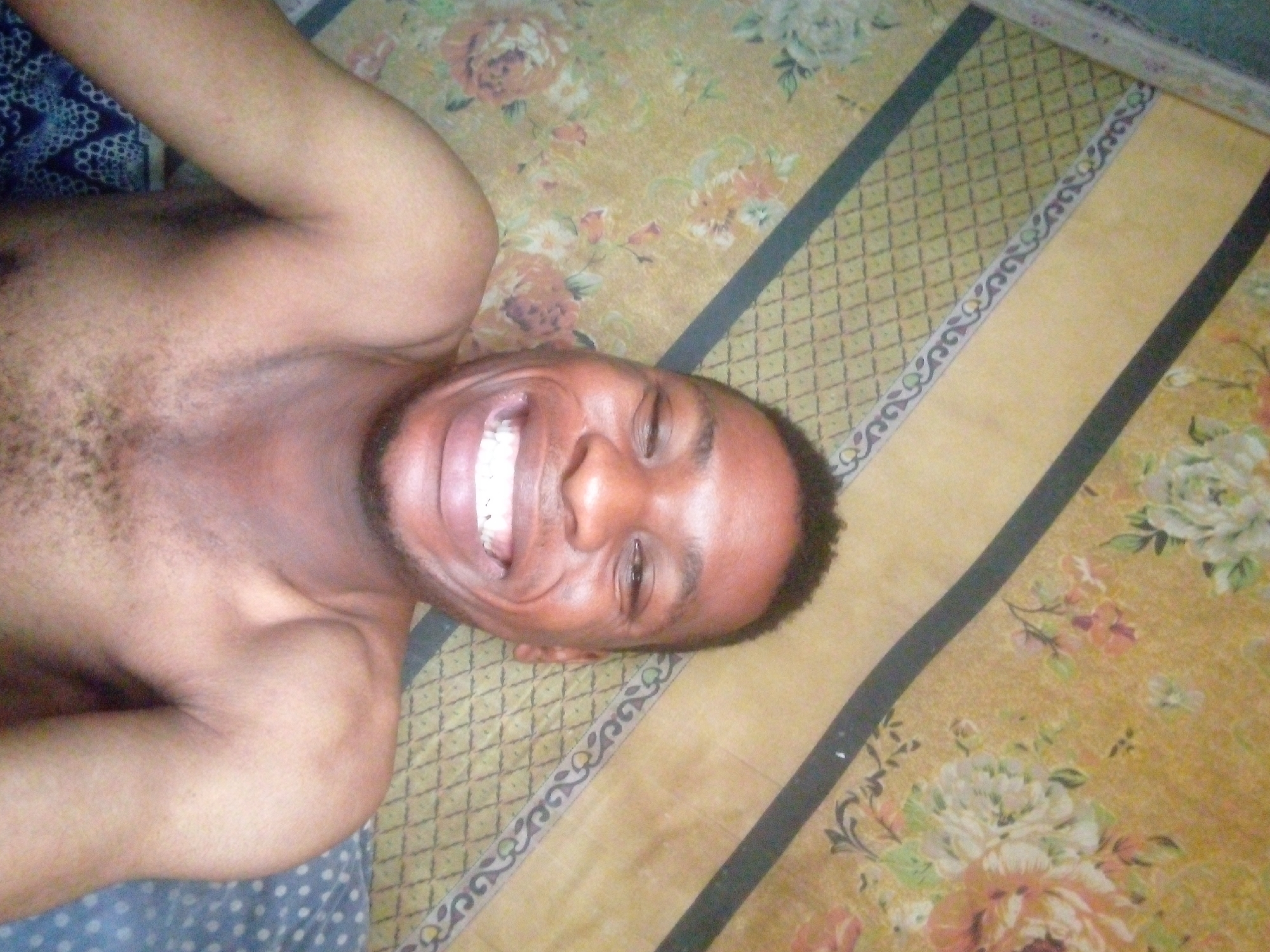 A man is lying on his back on a floral-patterned bedspread with striped accents. He is shirtless and smiling broadly. The location appears to be indoors. The city is Unknown, Unknown.