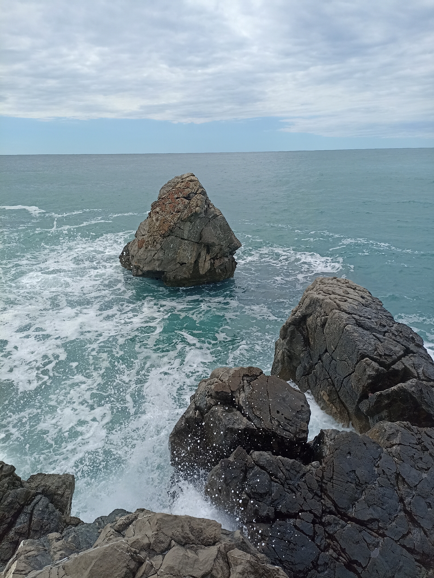 The image shows a rocky coastline in Čanj, Montenegro. Large, dark gray rocks occupy the foreground and right side of the frame, with seawater crashing against them and creating white foam. In the middle ground, another, lighter-colored rock formation rises from the sea. The sea extends to the horizon under a cloudy sky, with shades of gray and some areas of blue visible. The overall impression is one of a rugged, natural coastline with dynamic wave action. No people are visible.