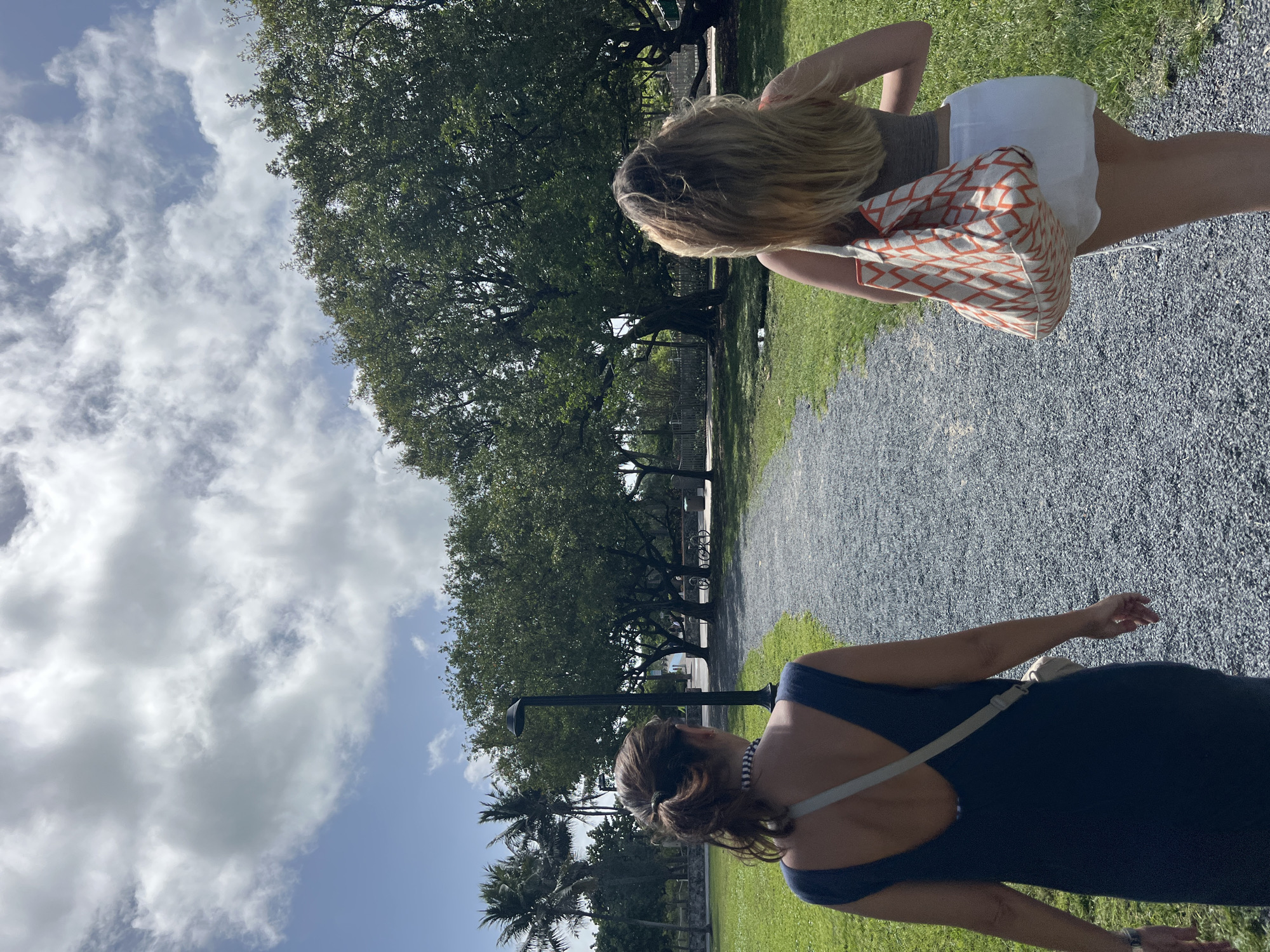 Two women are walking along a gravel path in a park. The woman in the foreground is wearing a navy blue dress with a light-colored strap across her back. The woman in the background is wearing a gray top and white shorts, and carrying a patterned bag. The park has large trees and green grass. There is a lamp post and parked bikes in the background. The sky is blue with white clouds. The location appears to be in a park in Miami Beach.