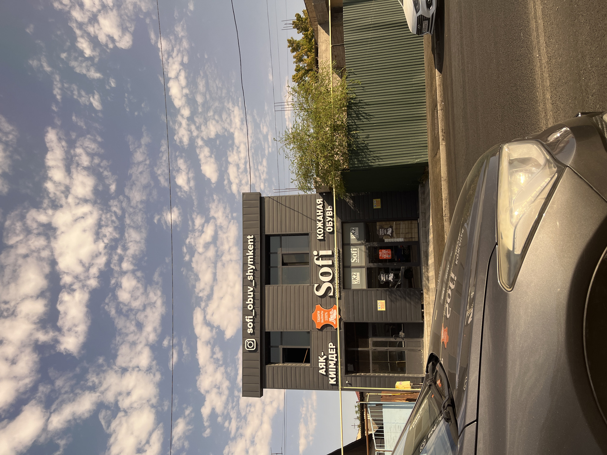 The image shows a two-story dark gray building with a sign that reads "sofi_obuv_shymkent" and an Instagram logo. The building has the word "Sofi" in large white letters, and text in Cyrillic on either side. Above the entrance, on the left side, is the Cyrillic text "АЯК - КИИМДЕР", and on the right is the Cyrillic text "КОЖАНАЯ ОБУВЬ". There is an orange leather-shaped logo with "ТАБИГИ БАЙЛАМ 100%" printed on it. Below the main sign, on the ground floor, there are multiple windows and doors. The building is situated on a street in Shymkent, Kazakhstan, as indicated by the signage. In the foreground, the hood and headlight of a dark gray car are visible. A green corrugated metal fence runs along the side of the building, behind which trees and parts of other structures are visible. A white car with a license plate is parked further down the street. The sky above is blue with scattered white clouds.