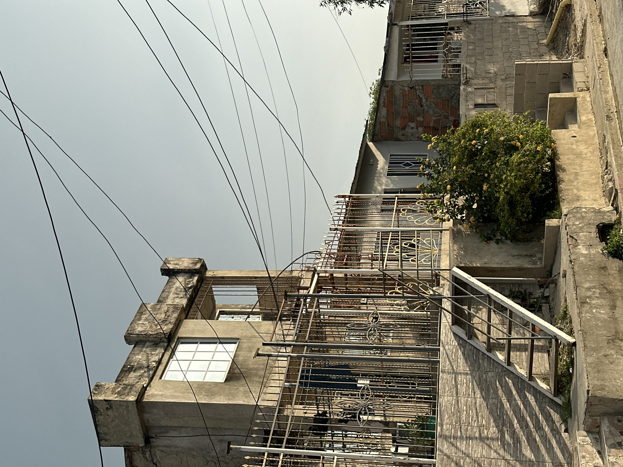 The image depicts a street scene in Perímetro Urbano Barranquilla, Colombia. The foreground shows a sidewalk with a curb and street, transitioning into a concrete area with overgrown grass and a small set of stairs. To the left, a textured tiled wall leads up to a metal staircase with white steps and handrails, which ascends to a wrought-iron fence with ornate scrollwork. Behind the fence, a two-story building with a rough concrete facade and a flat roof is visible. The lower portion of this building has a large window with a grid pattern, and above it, a smaller window with a similar grid. A spiral staircase is visible on the right side of this building, leading up to a balcony with a metal railing.
To the right of the main building, a smaller, single-story structure is partially visible. This structure appears to be under construction or renovation, with exposed brickwork on one section and concrete block walls on another. It also has a wrought-iron fence and a window with a grid. A lush green bush with yellow flowers is growing in front of the single-story building, near the base of the metal staircase. Numerous black electrical wires crisscross the sky, which is a pale, hazy blue, suggesting either early morning or late afternoon light. The overall impression is of a moderately weathered urban environment, characterized by a mix of established and partially completed structures, and abundant overhead wiring. No people are present in the image.