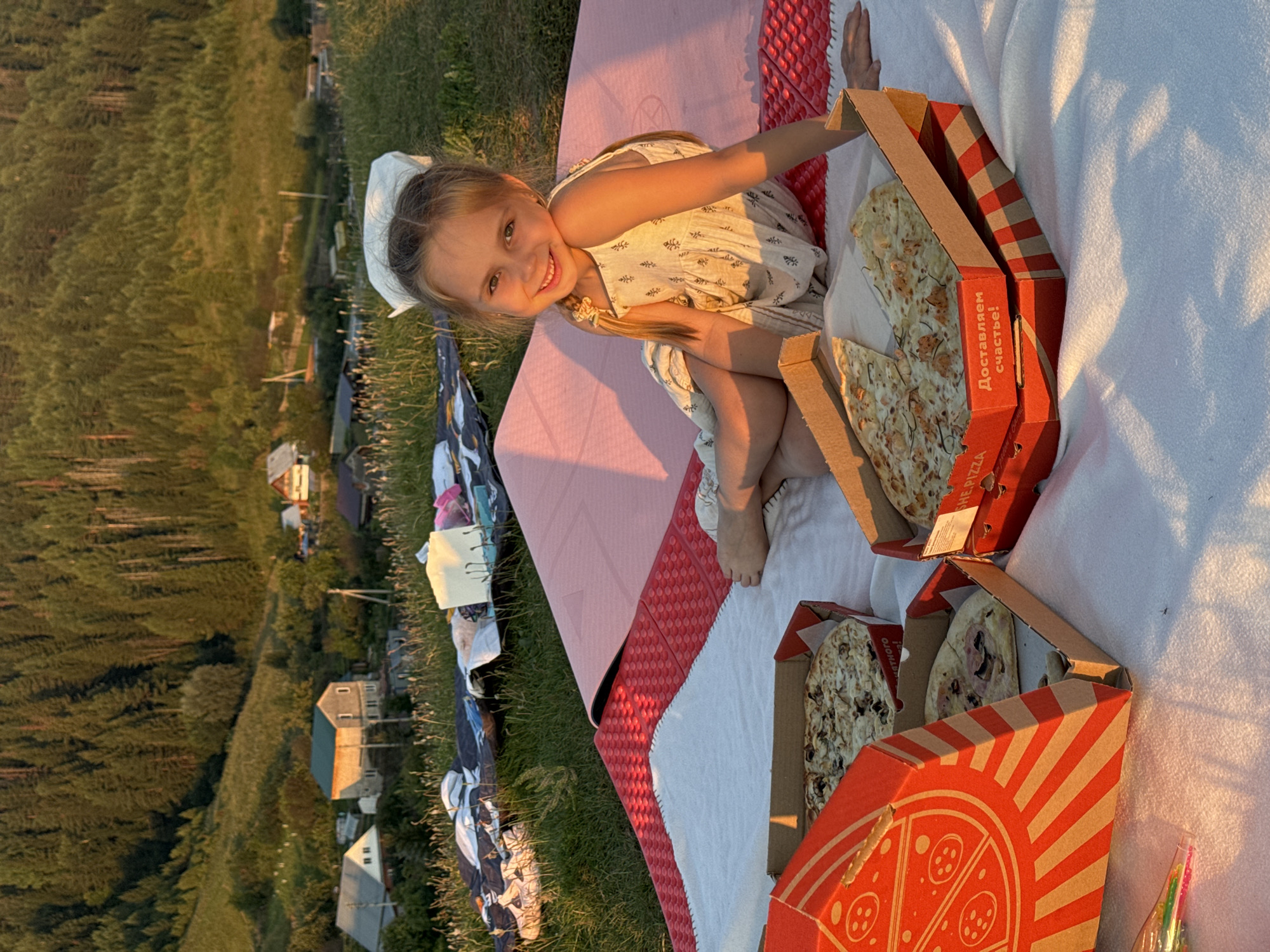 A young Caucasian girl with blonde hair in a ponytail and a white bow is sitting on a blanket outdoors, smiling at the camera. She is wearing a light-colored dress with a subtle pattern. In front of her, on the blanket, are several pizza boxes, two of which are open, revealing pizzas. The girl is positioned on a pink mat, which is on top of a red mat with a textured surface. In the background, a grassy slope leads up to a dense forest. Several houses and structures are visible nestled among trees on the hillside. The scene appears to be in a rural area, possibly on the outskirts of Ольховка, Russia, given the architecture and natural landscape. The lighting suggests it is late afternoon or early evening, with warm sunlight casting long shadows.