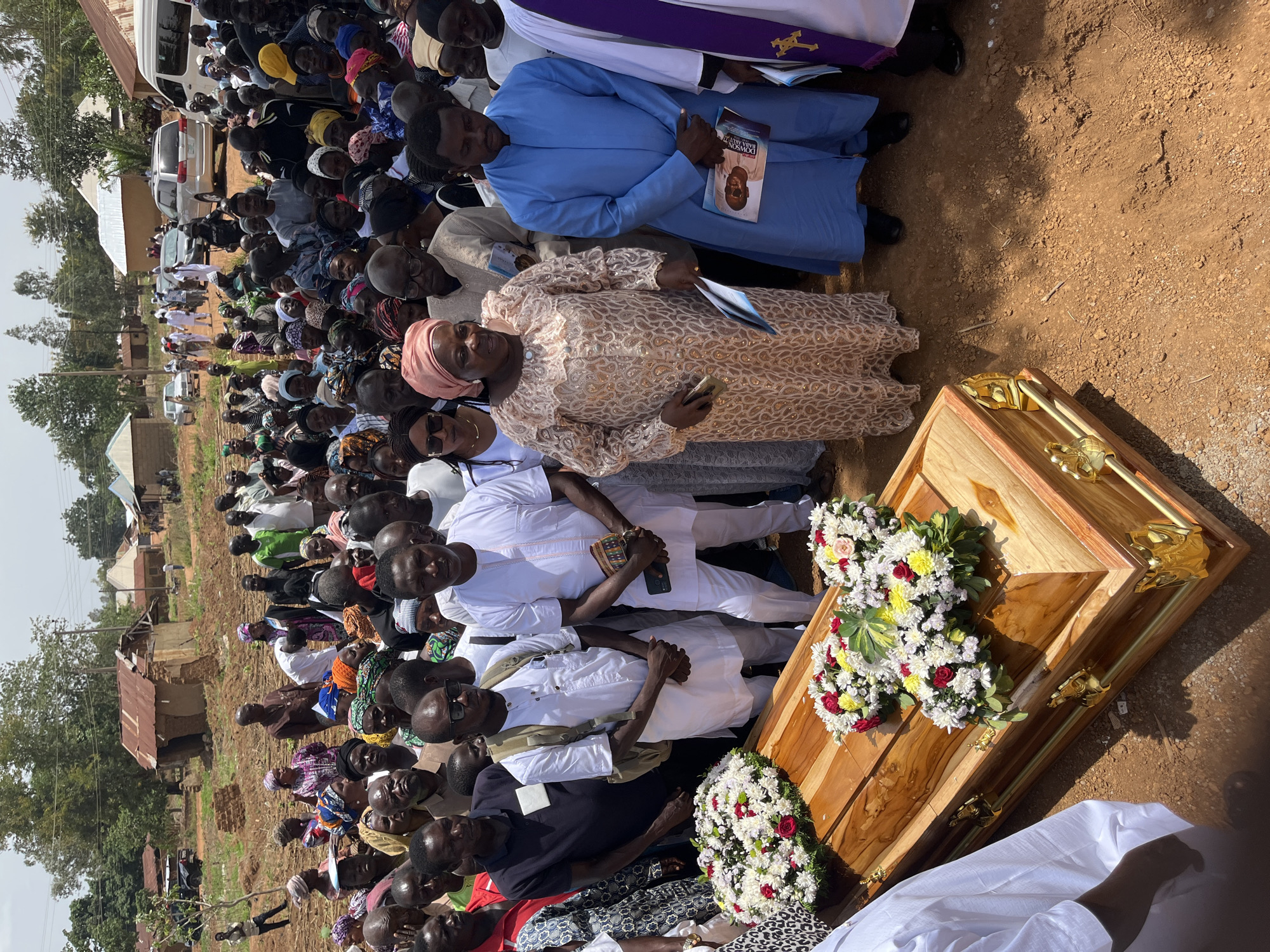 A gathering of people are standing outside next to a wooden casket adorned with white, red, and yellow floral arrangements. The location appears to be Dogon Dinia, Nigeria based on contextual information. The area is open, with a reddish-brown dirt surface. People are dressed in a variety of clothing, including traditional attire, casual wear, and religious vestments. Some people are holding books or pamphlets. The overall atmosphere appears somber, suggesting a funeral or memorial service.