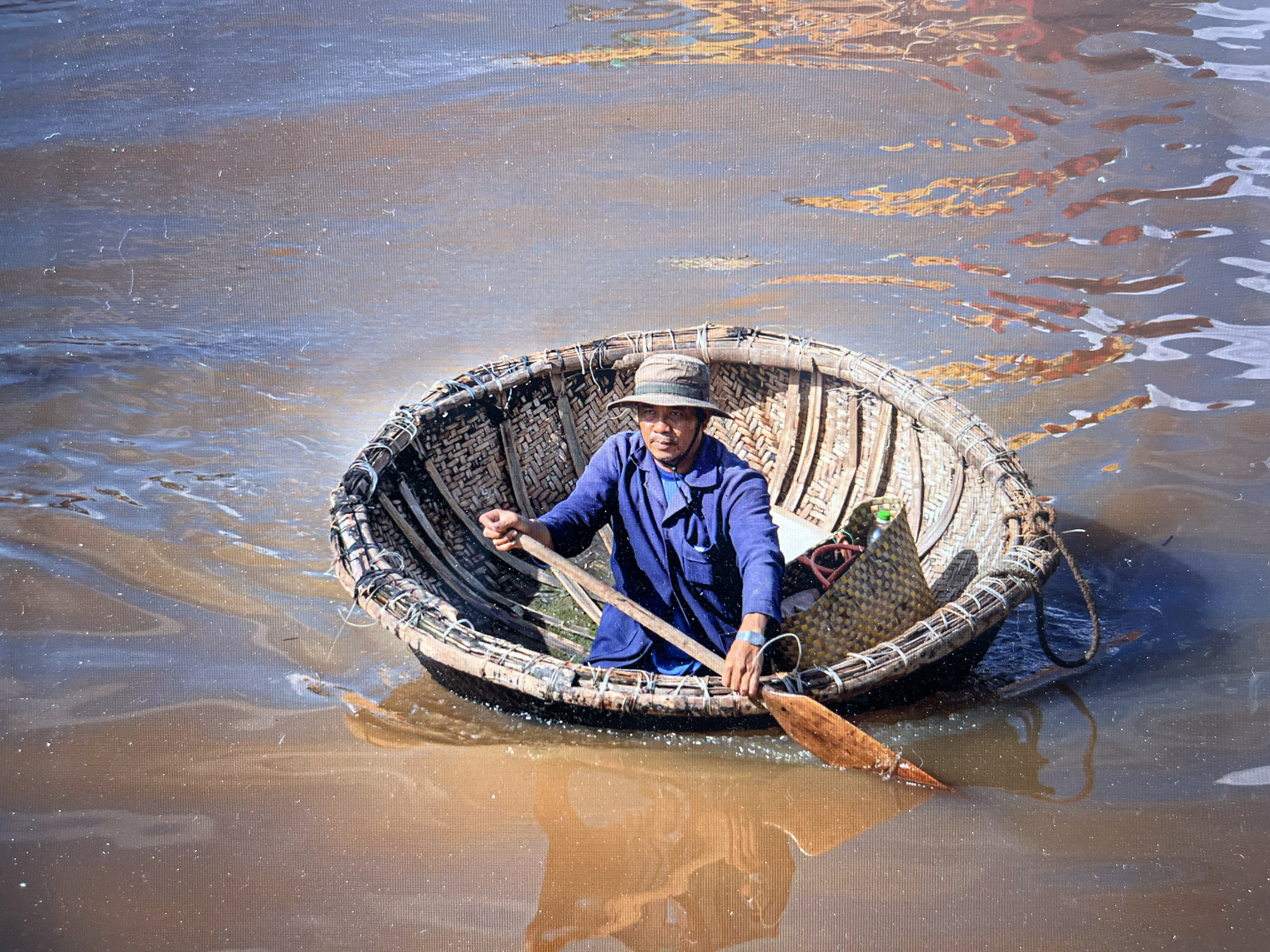 A man in a blue jacket and hat is seated in a coracle-like boat, using a paddle in the water. The boat is made of woven material and has a woven basket inside. The water is murky brown with blue areas and reflections. The image appears to be taken on a canvas or textured surface. The location is Bac Nha Trang Ward, Vietnam.