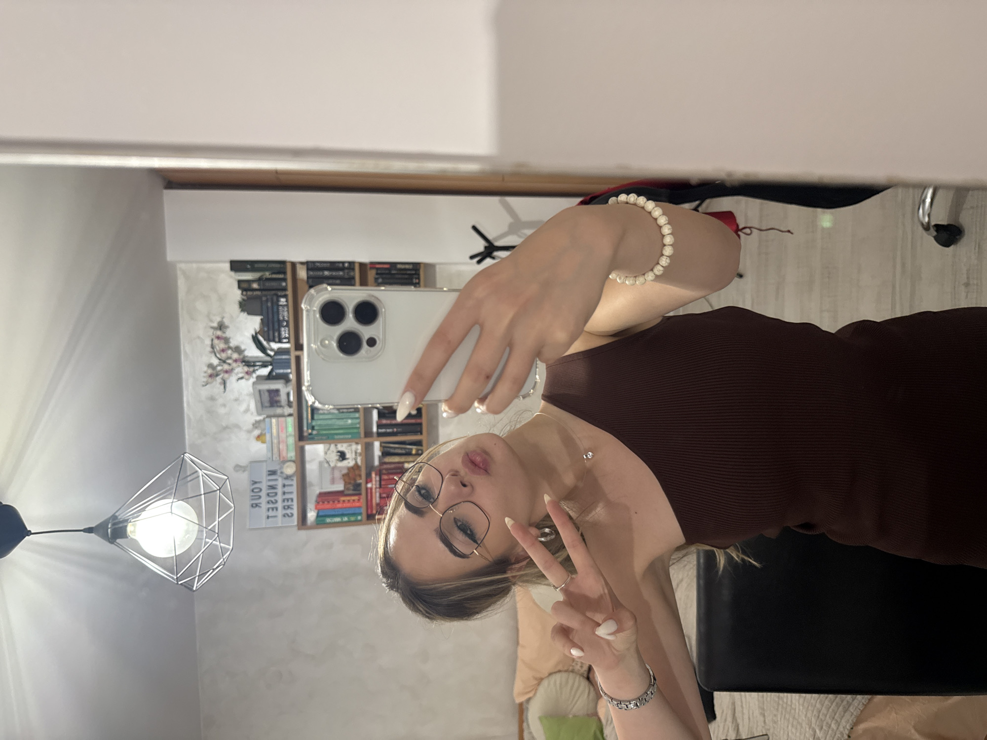 The image shows a woman taking a selfie in a mirror. She is wearing glasses and a brown tank top, and holding a smartphone. She is making a peace sign with her fingers. Behind her, there is a bookshelf filled with books, a lamp, and a sign that says "Your Mindset Matters". The location appears to be an interior space, possibly a home. There is no information about location in Blagoevgrad, Bulgaria, that is discernable from the image.
