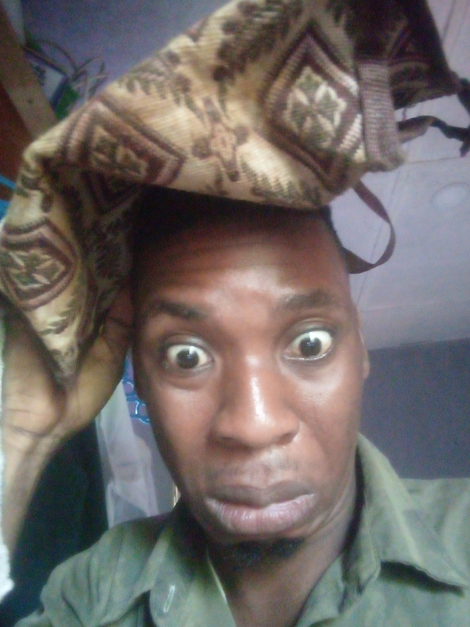 A person is taking a close-up selfie with a patterned fabric or bag draped over the top of their head. They are looking directly at the camera with a slightly surprised or exaggerated expression, their eyes wide. The person has dark skin, short hair, and is wearing a green collared shirt. The background is blurry and out of focus. The location is Unknown, Unknown.