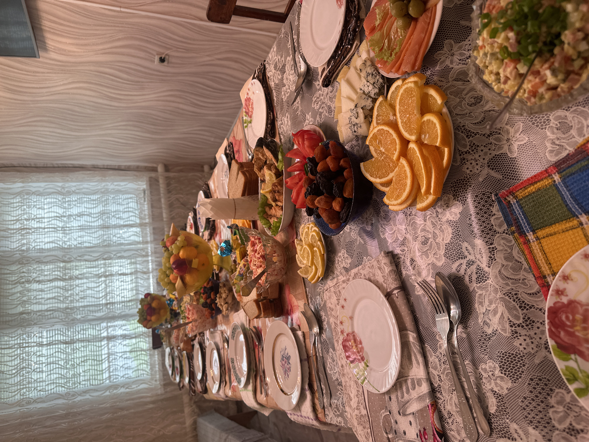 A table laden with food is positioned in a room, likely a dining area, in Коноваловка, Russia. The table is covered with a lace tablecloth. On the table, there are plates, silverware, fruit arrangements, salads, slices of orange, blue cheese, cured fish, dried fruits, and bread. A window with a curtain is visible in the background. The ceiling has a textured, wavy pattern.