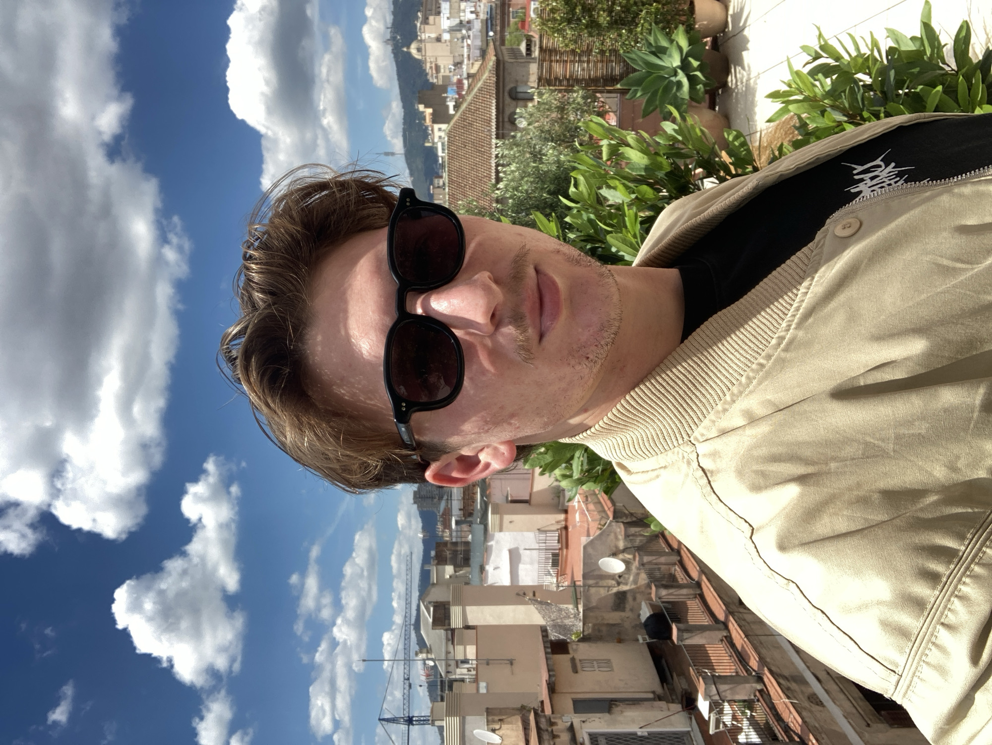 A medium-complex horizontal indoor shot captures a man wearing sunglasses and a light-colored jacket, with a cityscape and a bright sky in the background. The man appears to be on a rooftop or elevated location, as evidenced by the surrounding buildings. The cityscape in the background features buildings of varying heights and architectural styles, with some visible antennas and satellite dishes. There's a hint of green foliage interspersed among the buildings, possibly indicating trees or plants on balconies or terraces. In the distance, there are mountains visible. The sky is blue with puffy white clouds. Based on the architectural style and general layout of the cityscape, the location appears to be Barcelona, Spain.