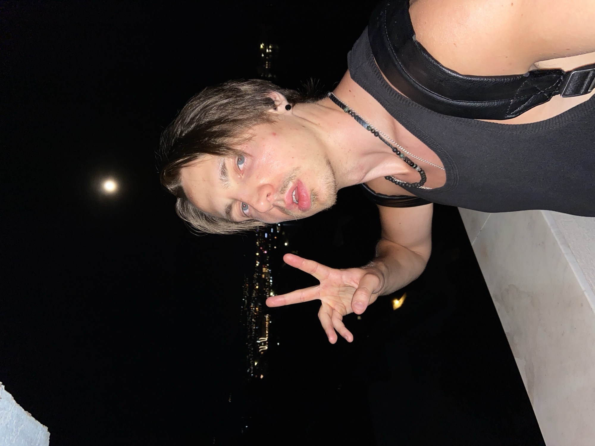 A young man with dark blonde hair is leaning over a ledge, making a hand gesture against the dark night sky. He is wearing a black tank top with a leather harness and a beaded necklace. The bright moon is visible in the upper left corner. In the background, blurred lights suggest a cityscape, identifying the location as Šušanj, Montenegro. The ledge appears to be made of light-colored stone.