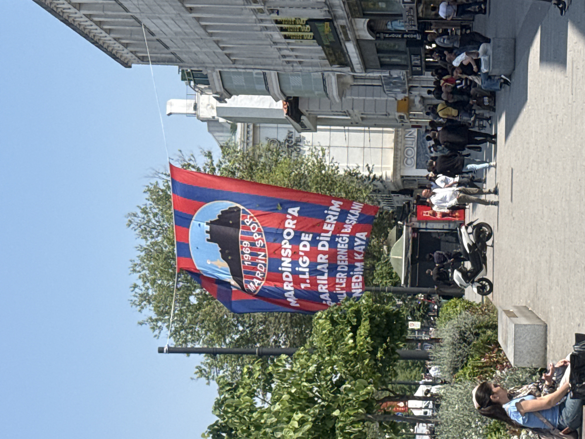 The image depicts a vibrant street scene in an urban environment, possibly in Turkey. In the foreground, a flag hangs prominently, bearing the name "MARDINSPOR" and the year "1969" along with the message wishing "Parilar" success in the first league and the name Nedim Kaya. The flag is red and blue striped with a emblem featuring a building silhouette. In the background, there is a busy pedestrian area with people walking, sitting, and standing. A motorcycle is parked on the side of the street. Buildings with signs such as "COLIN" and "Bijoux Bacos" and "STORY HOTEL" are visible, suggesting a commercial district. The sky is clear and blue, and some greenery is present, including trees and bushes.