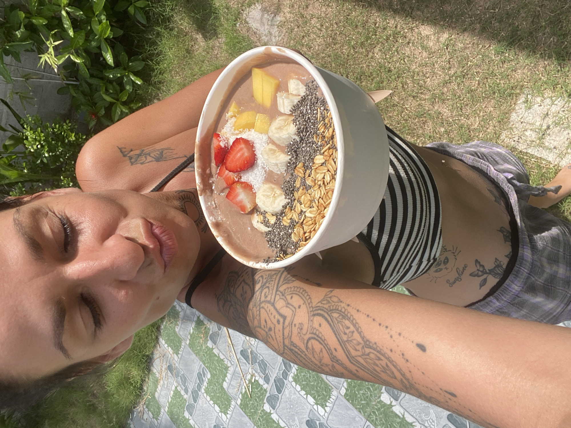 A woman in a black and white striped tank top and plaid shorts is holding a smoothie bowl with fruit and granola. She is outdoors, with grass and foliage in the background. Her arms and abdomen are covered in tattoos. The sun is shining brightly. The scene appears to be in Mũi Né, Vietnam.
