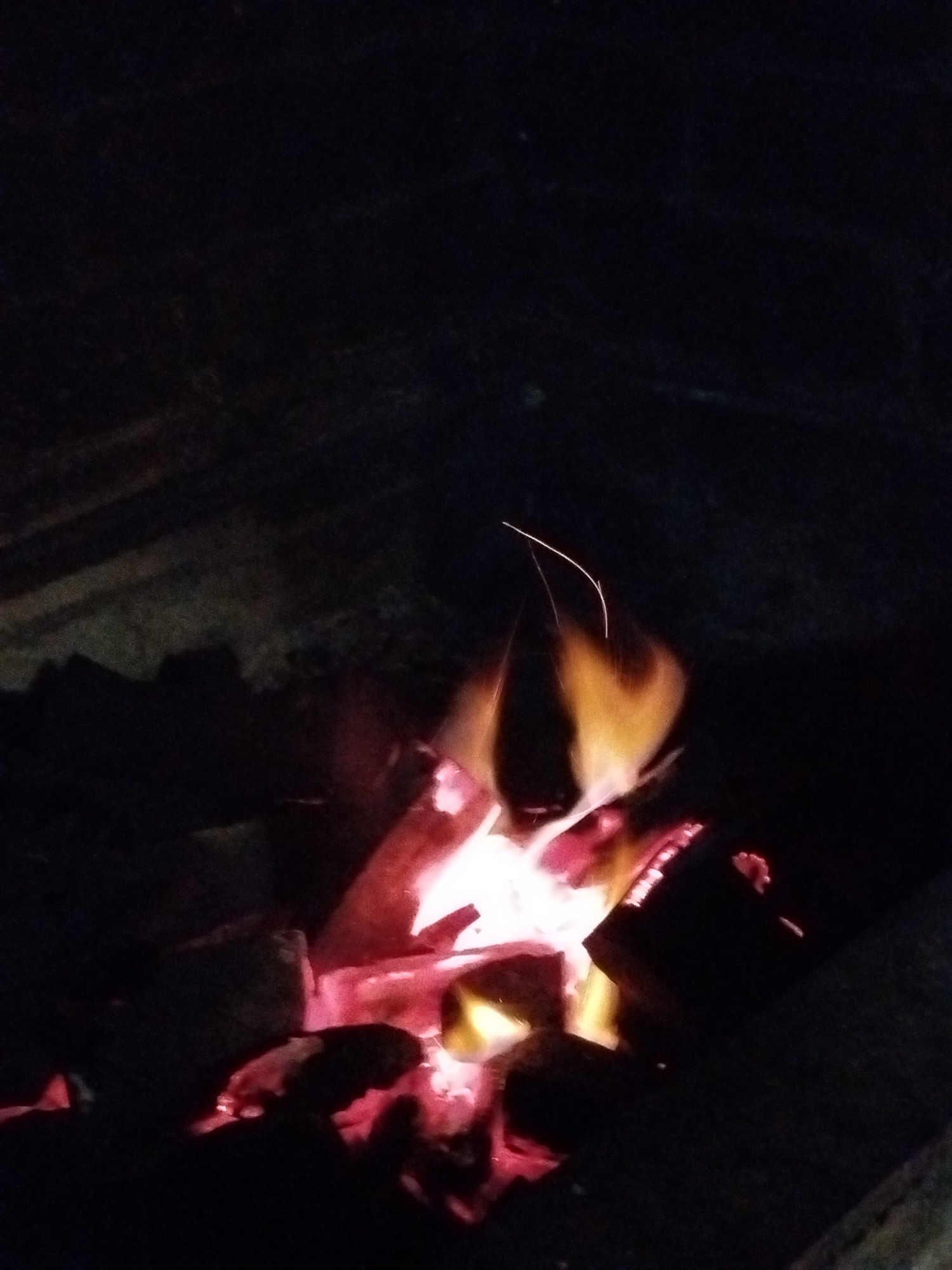 The image shows a close-up view of a fire pit at night. The fire is burning with vibrant orange and red flames, illuminating the surrounding charcoal and dark environment. There are embers glowing and sparks flying upwards, creating a dynamic visual. The background is predominantly dark, suggesting it is nighttime or a dimly lit area. The location is specified as Karaman 1-Yy, Russia. There are no people or specific objects that can be identified beyond the fire pit and its contents.