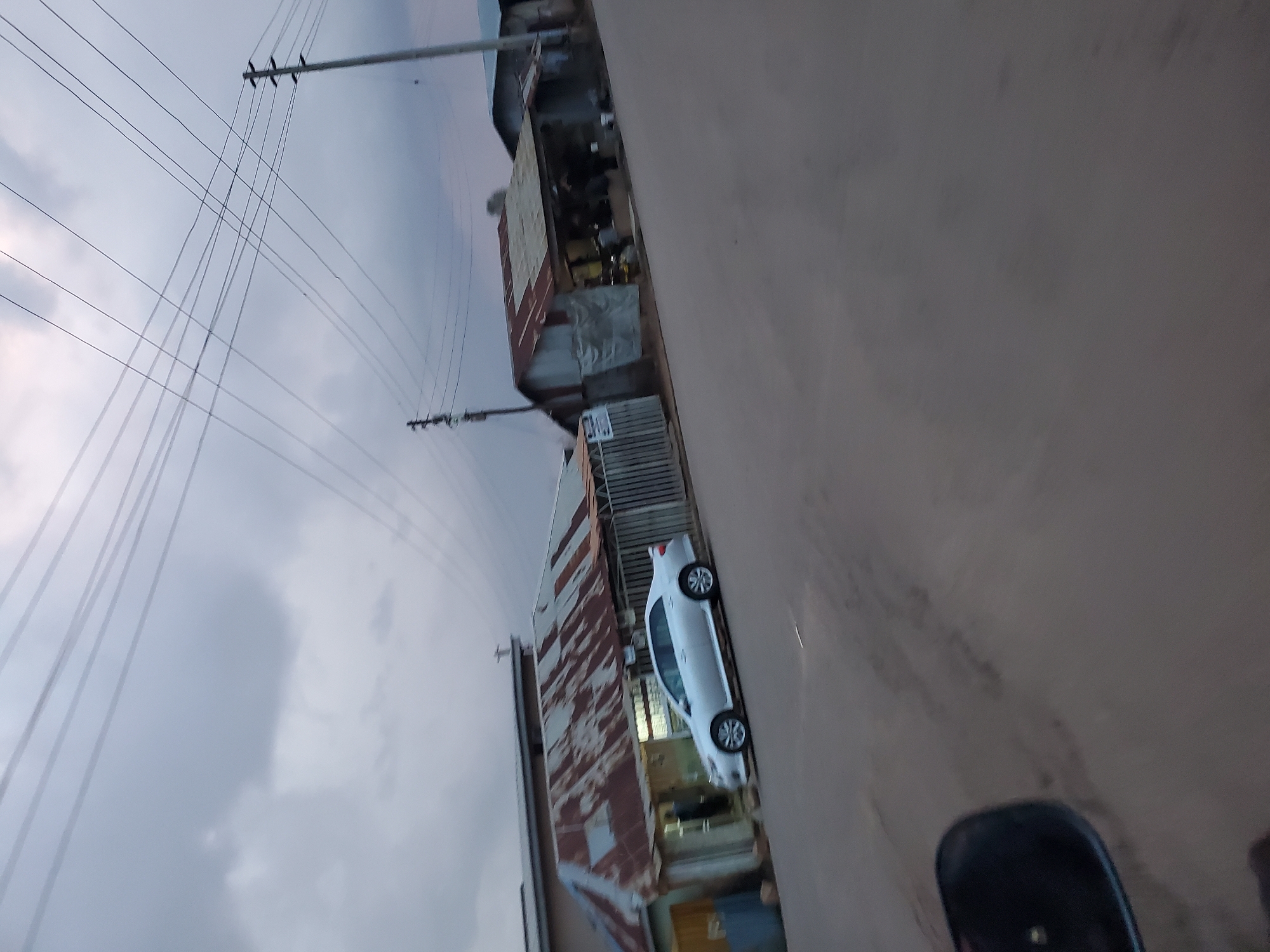 The image depicts a street scene in Jos, Nigeria, under an overcast sky. The foreground is dominated by a gray asphalt road, tilted to the right, with a portion of a black car side mirror visible in the lower left corner. In the mid-ground, a silver sedan is parked on the side of the road in front of a row of buildings. These buildings have corrugated metal roofs, some showing signs of rust and wear. The structures appear to be shops or small businesses, with openings that suggest merchandise might be displayed or services offered within. A metal fence runs along the front of some of the establishments. In the background, electrical wires crisscross the sky, supported by utility poles, indicating the presence of a power infrastructure. The overall atmosphere is somber, possibly due to the cloudy weather or the time of day.