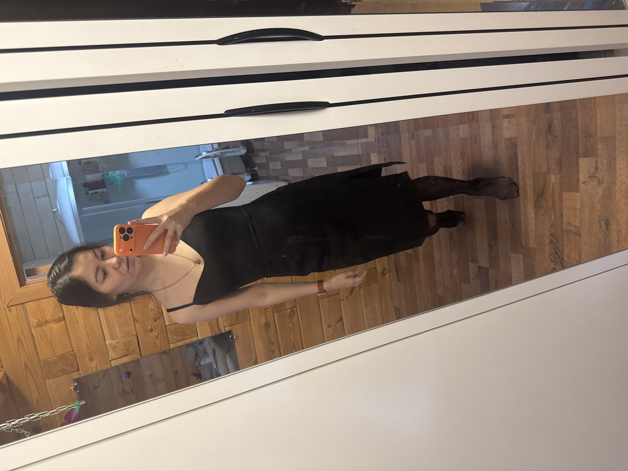 A young woman stands in front of a mirror, taking a selfie. She is wearing a black slip dress, dark pantyhose, and black shoes. The woman has dark hair and is wearing a necklace and a red watch. She is holding an orange smartphone in her right hand. The background appears to be a bathroom with wood-paneled walls and a tiled floor. In the reflection, a shower stall and various bottles are visible. The woman's pose suggests she is in a private setting, likely her home. The lighting is artificial, creating a warm, slightly dim atmosphere. The image is framed by a white closet door on the left and a tall, white wardrobe with black handles on the right.