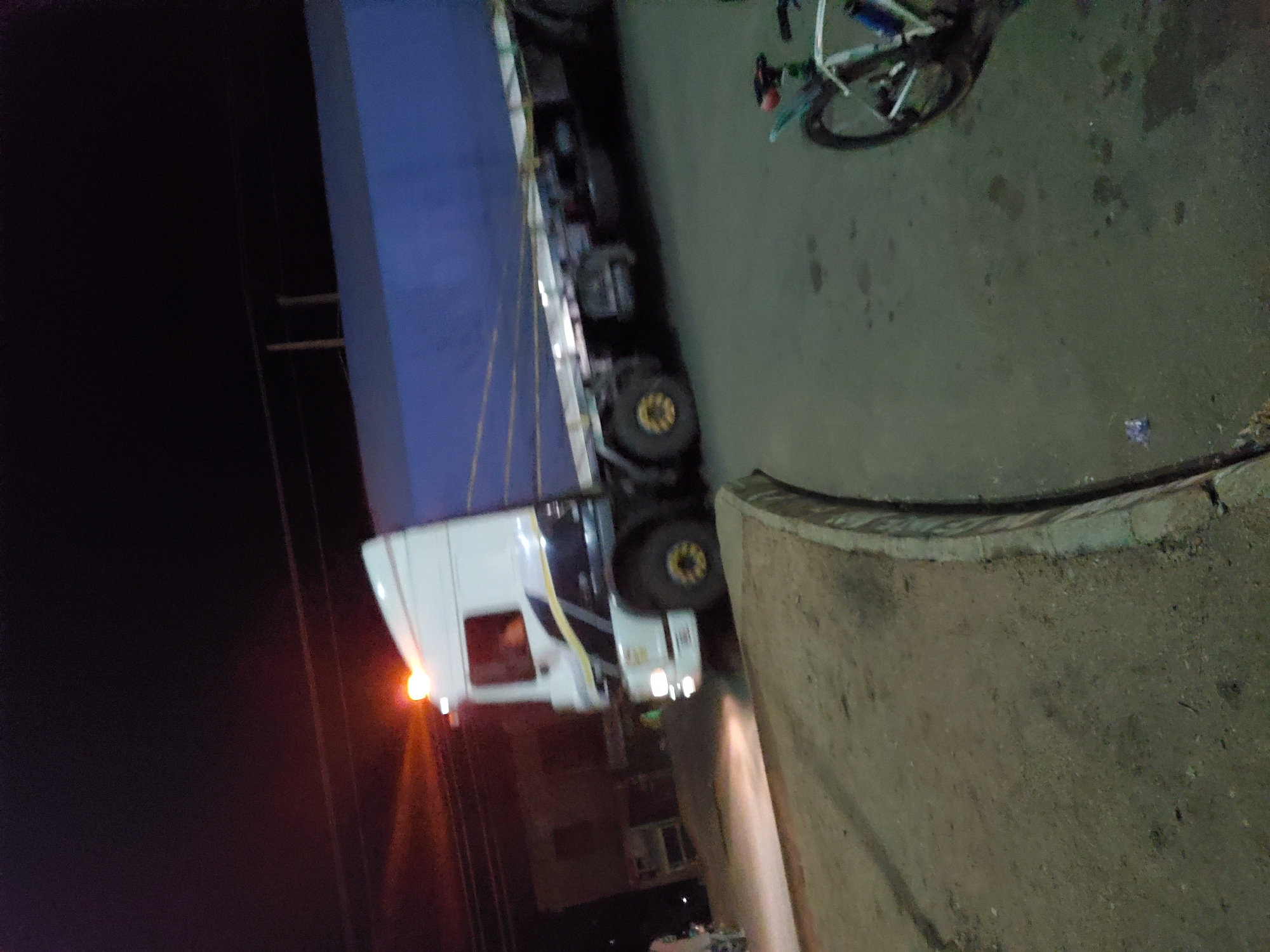 The image depicts a nighttime scene in Bukuru, Nigeria, centered on a large, white semi-truck with a blue tarp-covered trailer. The truck is positioned on a paved road, with its headlights and an amber flashing light on the cab's roof illuminated. Yellow detailing is visible on the truck's wheels and as a stripe on the lower cab. A blurry figure is discernible inside the cab. To the right, a portion of a bicycle, including its front wheel and frame, rests on the paved surface. The immediate foreground consists of a dirt shoulder adjacent to the paved road, separated by a worn concrete curb that arcs to the right. The paved road shows some dark, irregular stains or wet areas. In the left background, a structure is faintly visible in the darkness, with power lines overhead against the night sky.