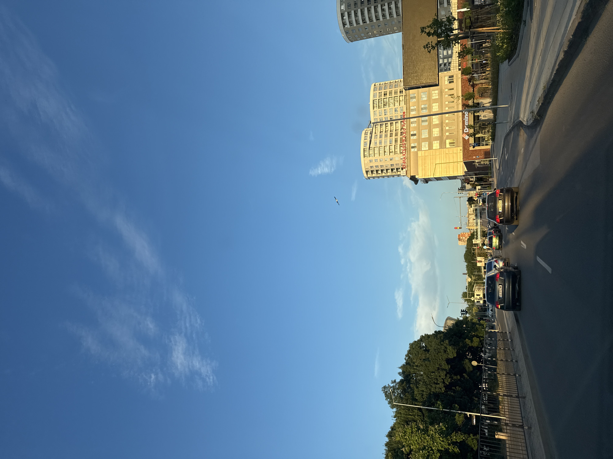 The image captures a street scene in Burgas, Bulgaria, likely during the late afternoon or early evening, given the warm, angled sunlight illuminating the buildings. The orientation of the image is unusual, as if the camera was held sideways.
The primary subjects are a clear blue sky with wispy white clouds, several modern multi-story buildings with many windows, a road with several cars, and a dense line of green trees. One of the buildings prominently features the "Domino's" logo in red letters. The architecture suggests an urban setting, with the buildings appearing to be residential or commercial complexes.
Cars are driving along the road, indicating typical city traffic. The lighting casts long shadows, hinting at the time of day. The overall atmosphere is peaceful and ordinary, depicting a regular day in the city. The presence of the trees suggests a well-maintained urban environment with green spaces. The buildings are varied in height and design, with one particularly tall, modern structure in the background. A solitary bird is visible in flight against the expansive sky.