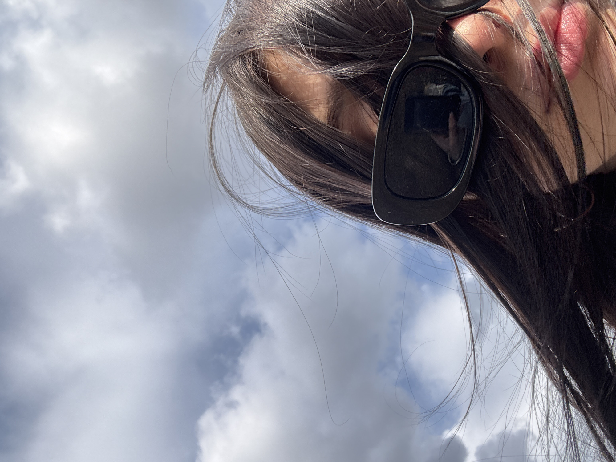 The image shows a person with long, dark hair and sunglasses, likely female, looking upwards towards a cloudy sky. The person is not fully visible, focusing on the area around the eyes, lips, and part of the hair and sunglasses. The location cannot be determined from the image. The reflection in the sunglasses lens does not provide any information about the location. No actions or interactions are discernible from the image.