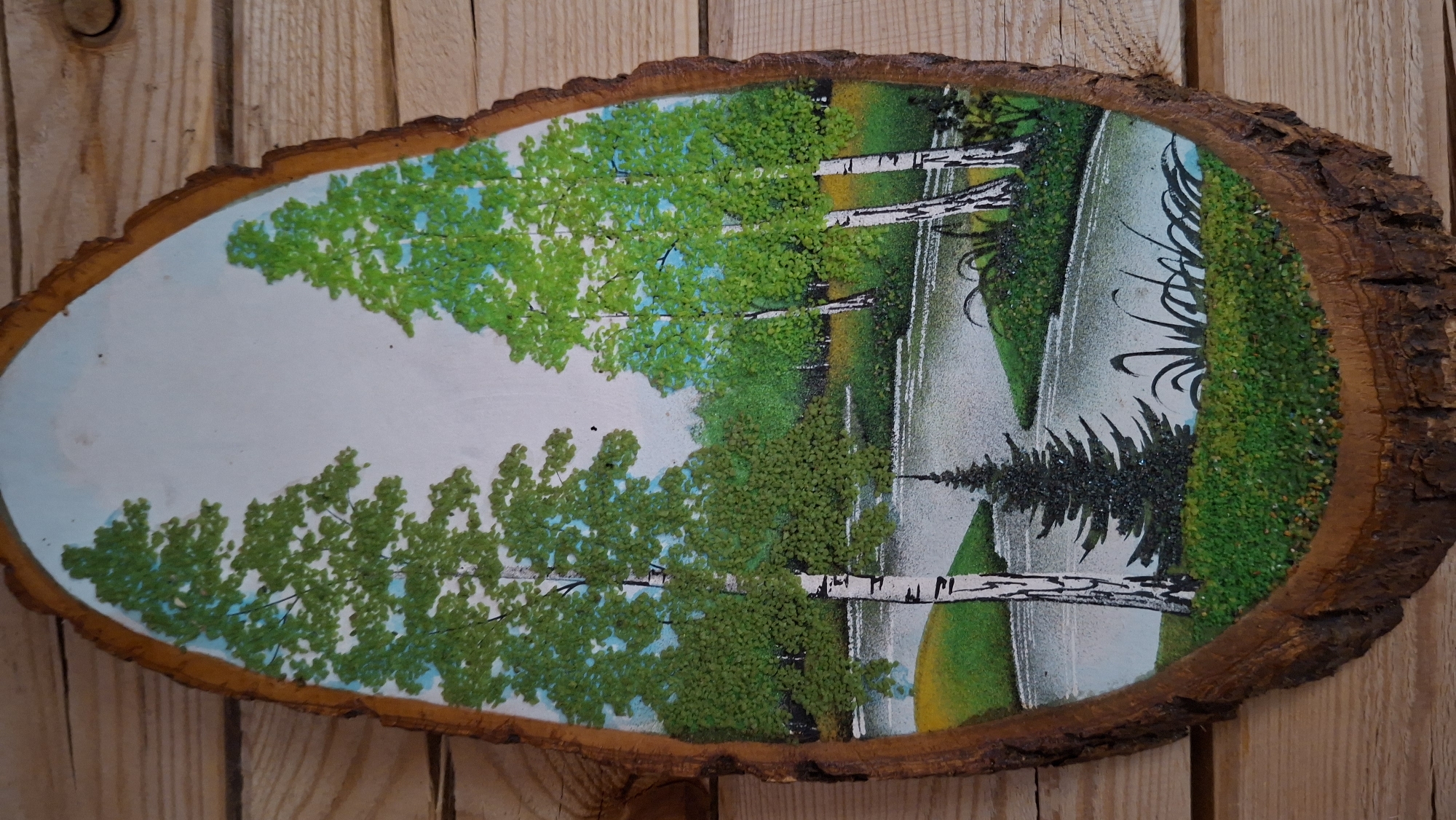 The image shows a cross-section of a tree trunk with a painting of a landscape on its surface. The landscape features birch trees with green foliage, reflected in what appears to be a body of water. A dark evergreen tree and other foliage are present along the edge of the water. The painting is on a light background, possibly representing the sky or a hazy atmosphere. The tree trunk cross-section is rough and textured with bark around the edge. The painting and tree trunk are mounted on a wooden surface, likely a wall or other display. The location is Большой Букор, Russia.