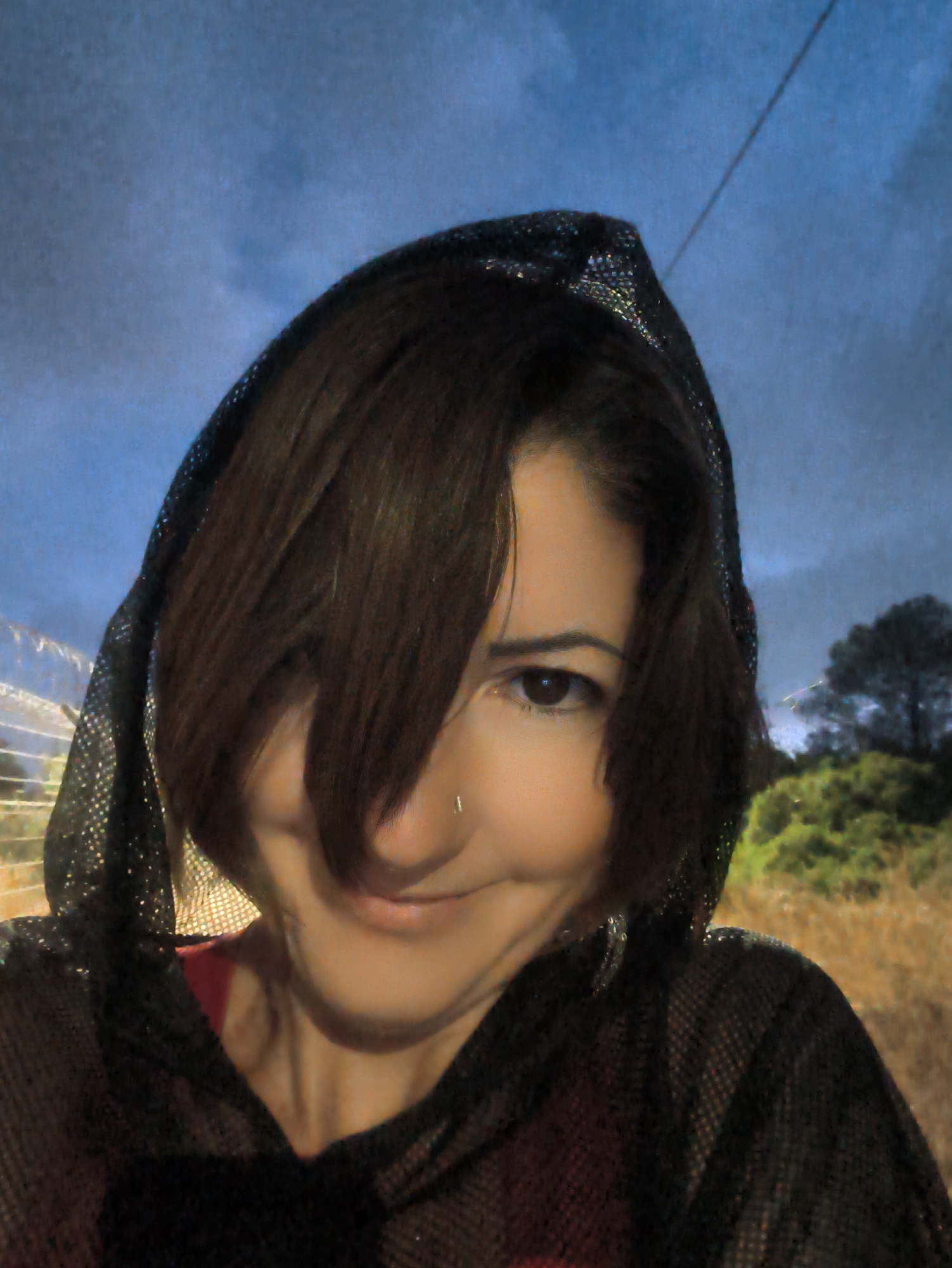 A medium shot, taken from a low angle, shows a woman wearing a black, sheer hooded garment. Her face is partially obscured by her dark brown hair, with one eye visible and a slight smile. She has a small nose ring. The background is a blurry landscape with a blue, cloudy sky. The scene appears to be outdoors, possibly in the city of Lachish Regional Council, Israel, given the type of vegetation and general environment depicted. A thin wire is visible in the upper right corner of the frame.