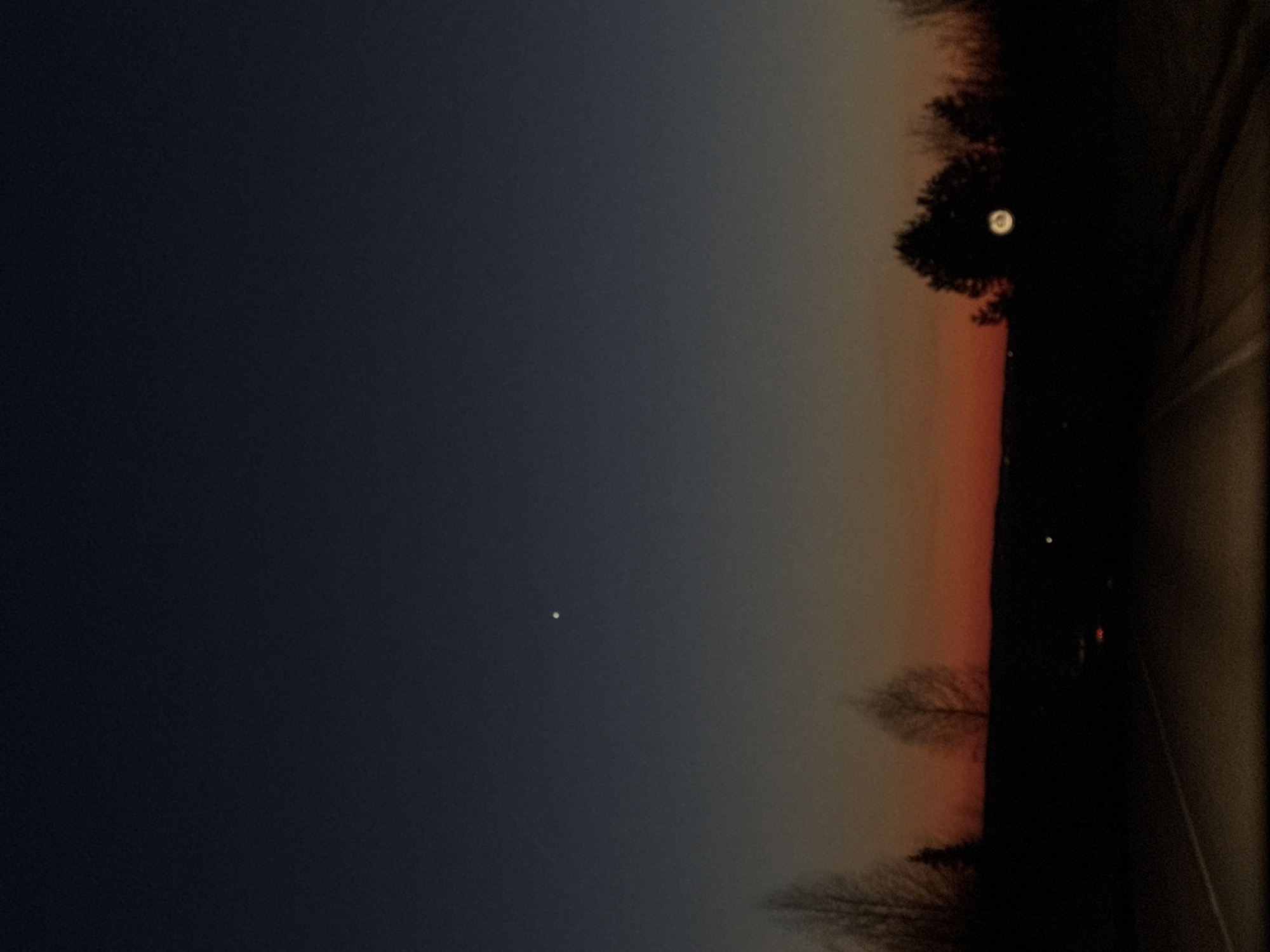 The image depicts a dark, partially illuminated scene likely taken during twilight. The sky transitions from deep blue on the left to orange/red on the right horizon, suggesting a sunrise or sunset. A single, small bright dot is visible in the sky, possibly a star or planet. Silhouetted trees line the horizon. On the right edge of the frame, a dark, undefined object is present, obscuring part of the view and suggesting a nearby surface, potentially the inside of a vehicle. There is a small, blurred light in the horizon. The image's location is speculated to be Garevaya, Russia.