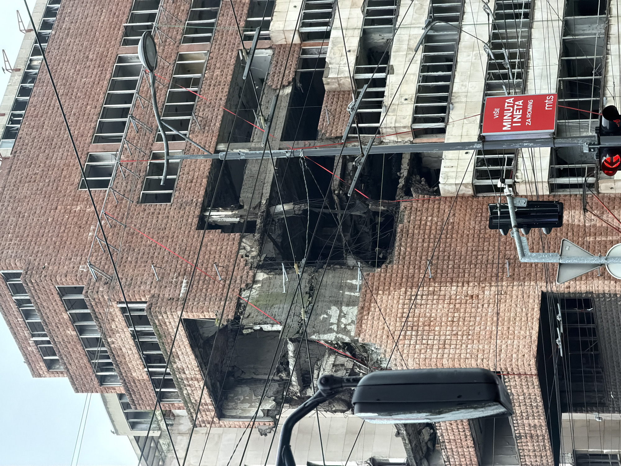 The image shows a bombed-out building in Belgrade, Serbia. The building's facade is damaged, with large holes and missing windows. A traffic light and street sign are visible in the foreground. The sign says "više minuta i neta za roming" and contains the MTS logo. Wires and cables are hanging from the building. The sky is overcast.