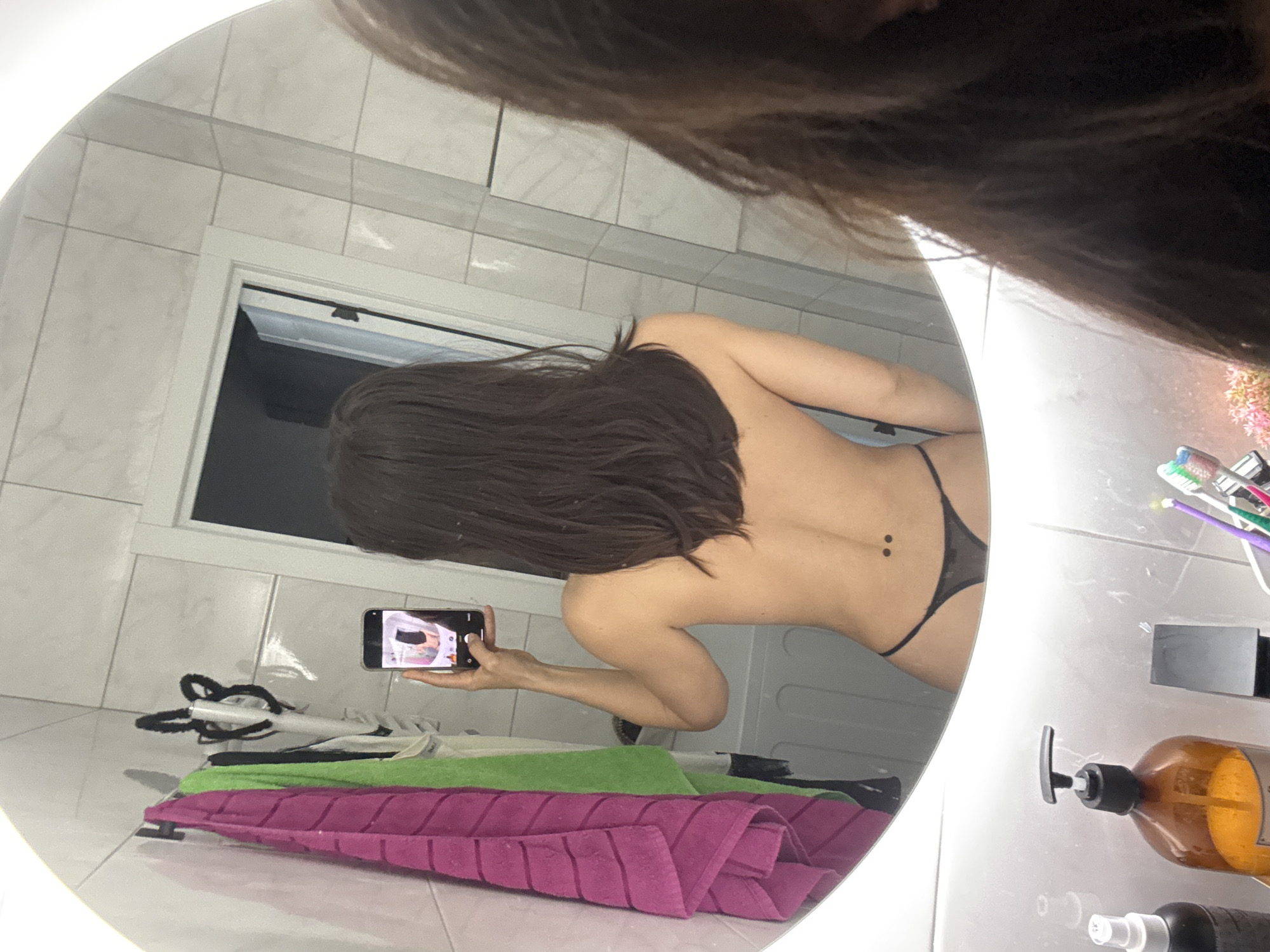 The image displays a person taking a mirror selfie in what appears to be a bathroom, located in Murino, Russia. The central element is a large, circular mirror with an illuminated rim, reflecting the back of a person with long, dark hair. The individual is wearing a dark thong, and a piercing is visible on their lower back.
The person is holding a black smartphone to capture the image, and the phone's screen itself shows a reflection of the person's front. The top of the person's head is largely obscured by their hair, which hangs into the foreground of the shot.
The setting features light-colored tiled walls and floor. A window in the background reveals a dark exterior, indicating it is likely nighttime or evening. On the floor below the mirror, folded towels, one magenta and one green, are visible, along with a white object that might be an appliance or part of a laundry setup. To the right of the mirror, a counter holds various bathroom items including a clear cup with multi-colored toothbrushes, a dark brown pump bottle (likely soap or lotion), and a black spray bottle. No discernible text is visible on any objects. The scene captures a private moment of self-documentation.