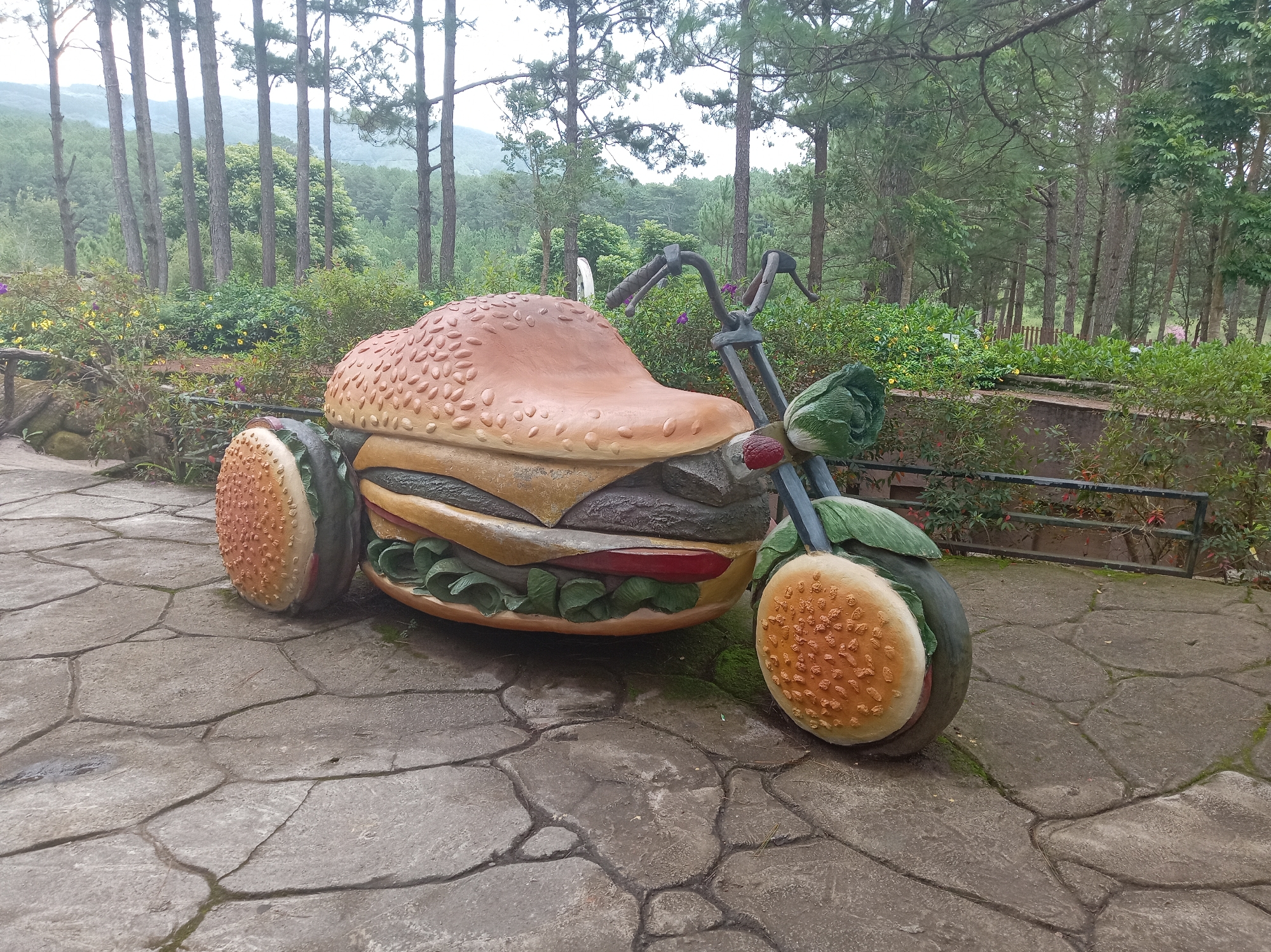 The image shows a quirky sculpture of a three-wheeled motorcycle designed to resemble a hamburger. The motorcycle's body is shaped like a bun, with layers of patty, cheese, lettuce, and tomato visible. The wheels are designed to look like burger buns, and the handlebars are topped with a lettuce-like material. The sculpture is situated on a paved pathway in a park-like setting with trees and greenery. The background features a dense forest with tall pine trees, suggesting a mountainous or highland area. This unique hamburger motorcycle sculpture is a known attraction in Da Lat, Vietnam, often found at various tourist spots like the Datanla Waterfall or in flower gardens, adding a touch of whimsical art to the natural scenery.