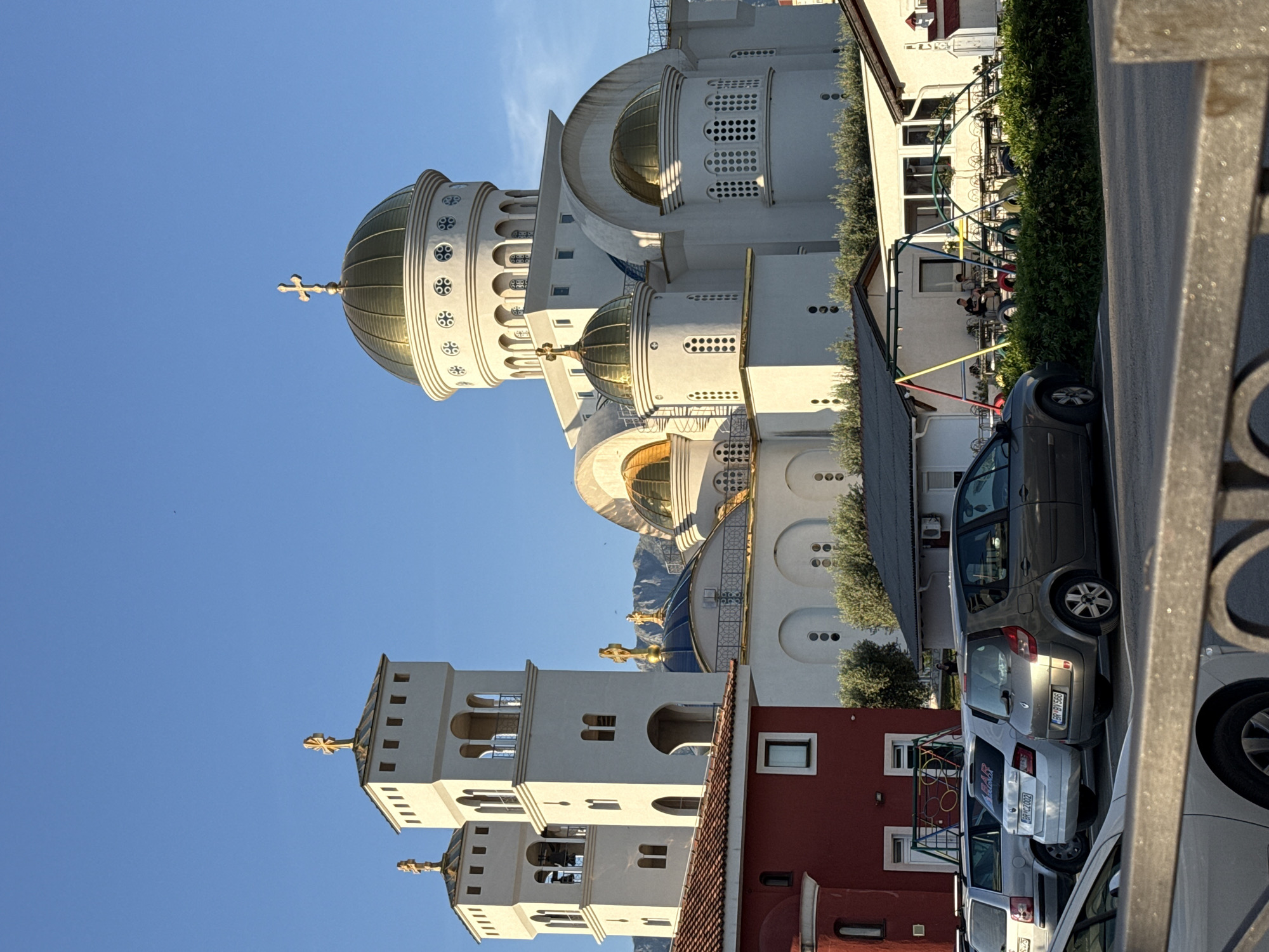 The image, taken in Bar, Montenegro, displays a prominent, ornate Orthodox church with multiple golden domes and crosses under a clear blue sky. The church features bright white facades with varied window styles, including arched and circular designs, and intricate circular patterns at the base of its larger domes. Two distinct bell towers, also white with golden crosses, stand to the left of the main church structure.
In the midground, residential buildings are visible, including a red-roofed building with white walls and a small window. Adjacent to this, a white building houses what appears to be an outdoor café or seating area, with a green hedge and a colorful, multi-arched structure that may be part of a playground or pergola. Several people are visible sitting at tables in this area.
The foreground and midground are occupied by parked cars. A dark grey minivan is prominently featured, along with two lighter grey cars. Visible license plates include "BP HY 566" and "BP CT 099", with another partial plate "BP 2002". The word "BAZAR" accompanied by a shopping cart icon is visible on the rear window of one light grey car.
The scene is set during daylight hours, indicated by the bright sun and clear, cloudless blue sky, which casts some shadows. The overall impression is a bustling urban or semi-urban environment where modern life intersects with striking religious architecture.