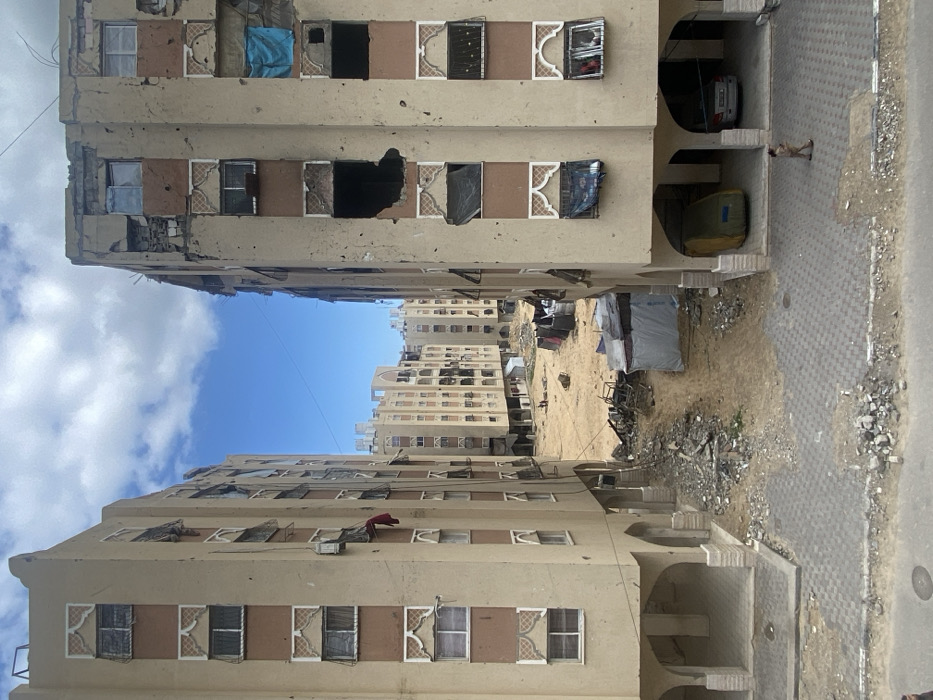 This image depicts a devastated urban landscape in Khan Yunis, Palestinian Territory, centered on a cleared, rubble-strewn area flanked by two severely damaged multi-story residential buildings. The buildings, beige with brown horizontal bands and decorative arched window frames, exhibit extensive structural trauma, including large, irregular holes exposing interiors, shattered and missing windows, and spalling on their facades. The building on the right has a large impact hole on its upper floors and another on a lower floor, with some windows boarded by blue tarpaulin. The building on the left shows numerous broken windows, with a red garment hanging from one.
The ground between these two structures consists of an unpaved dirt path heavily interspersed with construction debris, twisted metal, and broken concrete. A makeshift tent-like shelter, covered in light tarpaulin, stands on the right side of this open ground, near piles of dark material. In the background, beyond this cleared area, multiple similar multi-story residential buildings are visible, some appearing damaged, under a partly cloudy blue sky.
In the immediate foreground, a paved sidewalk composed of grey interlocking pavers is partially obscured by debris and dirt. A single individual, dressed in light-colored clothing, walks from right to left on this paved area in front of the right building's ground-level carports. Within these carports, a vehicle covered in a green/yellow tarpaulin and a white sedan are visible. Two small figures are faintly discernible in the distant open ground.