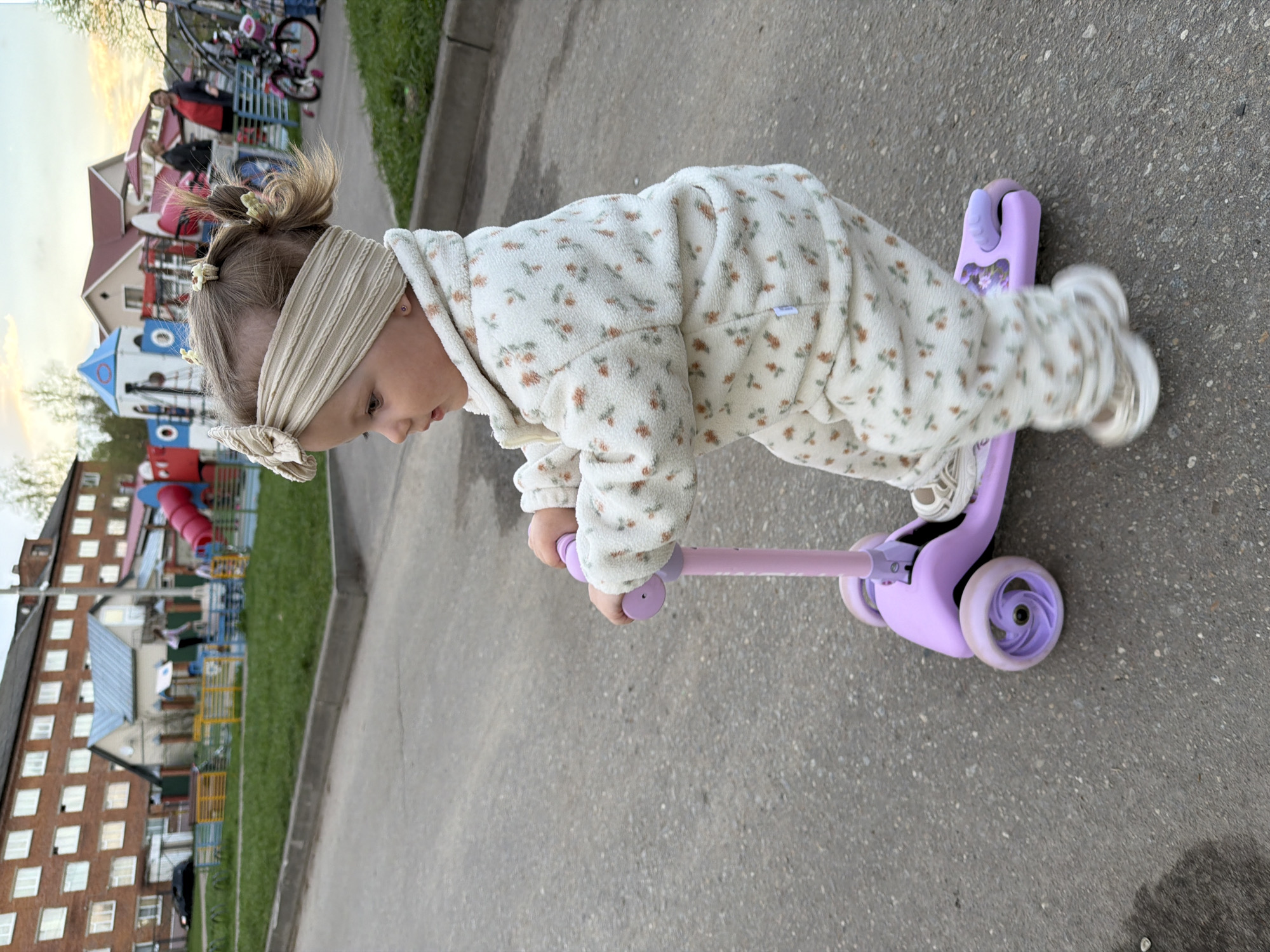 A young child in a floral-patterned jumpsuit rides a purple scooter on a paved surface. The child is facing left, gripping the scooter's handlebars, and wearing a headband. In the background, there's a grassy area, buildings including a brick apartment building, and a playground with slides and play structures. The location is likely Reshetnikovo, Russia, judging by the architectural style of the buildings.
