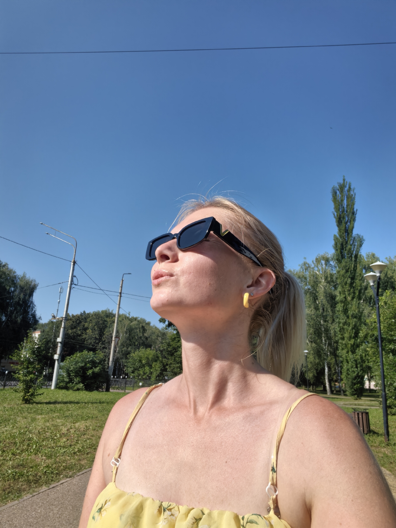 An outdoor, medium-shot, eye-level, selfie-style photograph captures a woman in Sterlitamak, Russia, on a sunny day. She is wearing black sunglasses, a yellow dress, and yellow hoop earrings. Her blonde hair is pulled back in a ponytail. The woman is looking upwards, with her lips puckered. In the background, there are trees, streetlights, and a clear blue sky with a power line. The woman is standing on a paved surface next to a grassy area.