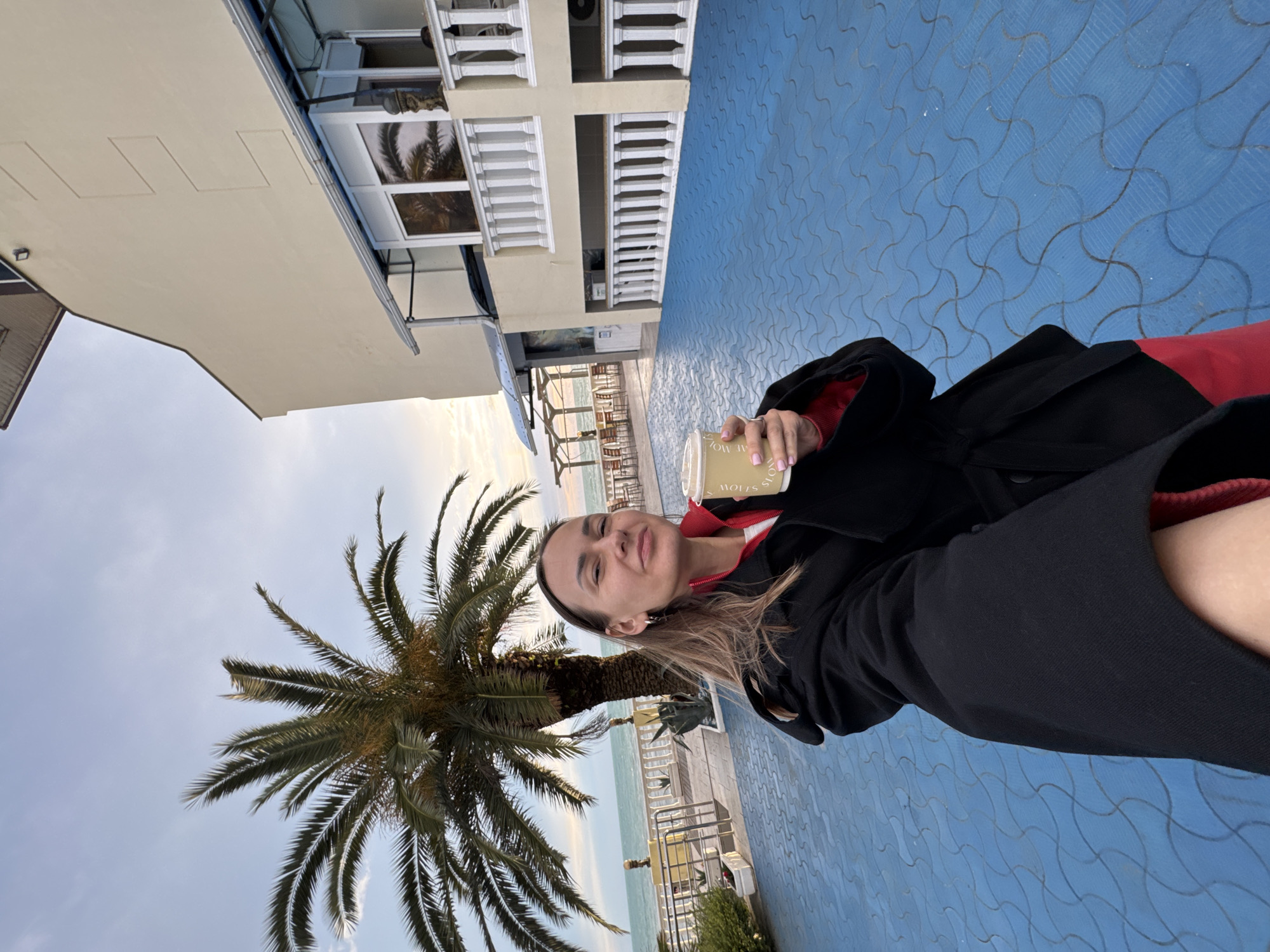 A woman stands on a blue-paved walkway, posing for a selfie. In her hand, she holds a cup. Behind her, there is a palm tree, the sea, and a building with white balconies. The scene is likely in Sochi, Russia, given the combination of the coastal setting, palm trees, and architecture.