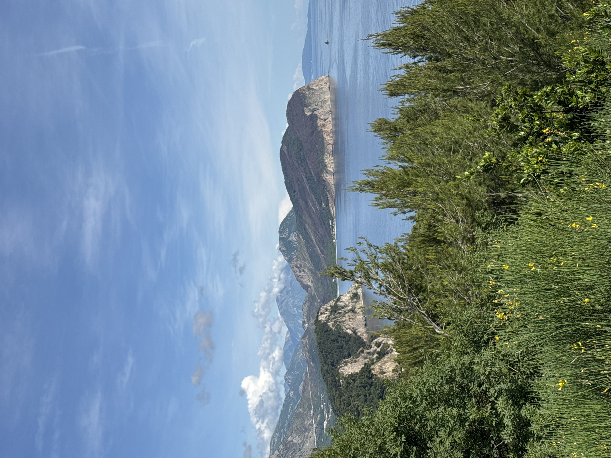 The image captures a coastal landscape in Mirište, Montenegro. A series of mountains stretch into the distance, partially obscured by clouds. A large, flat-topped rock formation is prominently situated in the water. Lush green vegetation, including trees and flowering bushes, fills the foreground. The sky is blue with wispy clouds. A small boat is visible in the water.